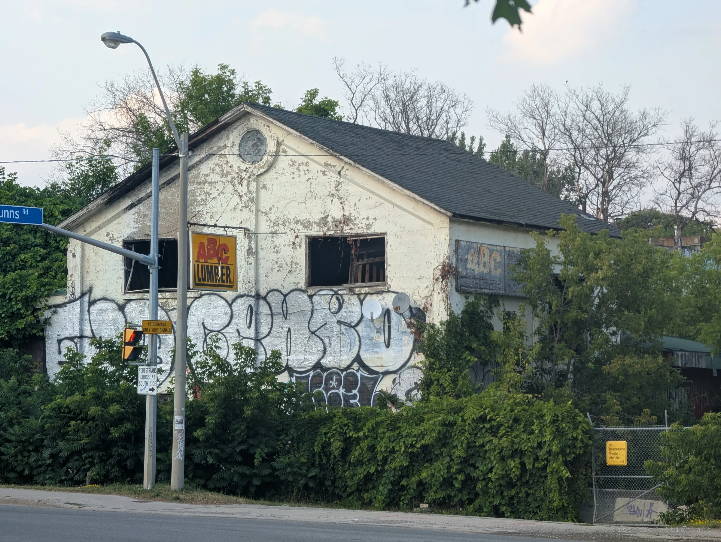 The peeling white gable of a derelict lumber yard building, its upper windows smashed out and its lower wall covered in chrome graffiti, half swallowed by vines beside a street corner.