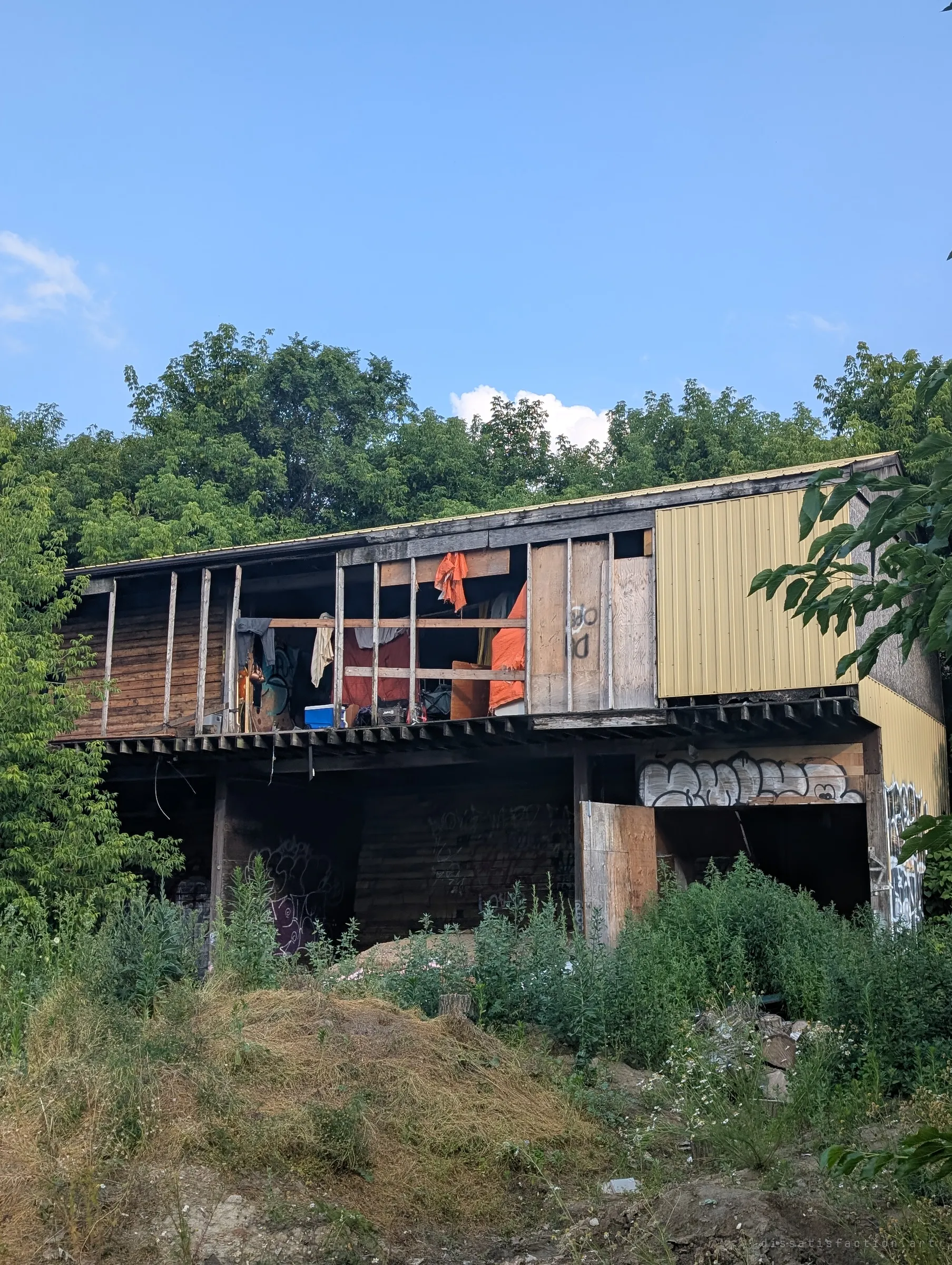 A two-storey open-fronted wooden shed seen from below an overgrown mound, its upper floor stripped to the studs and hung with blankets, tarps and a cooler where someone has made a home.