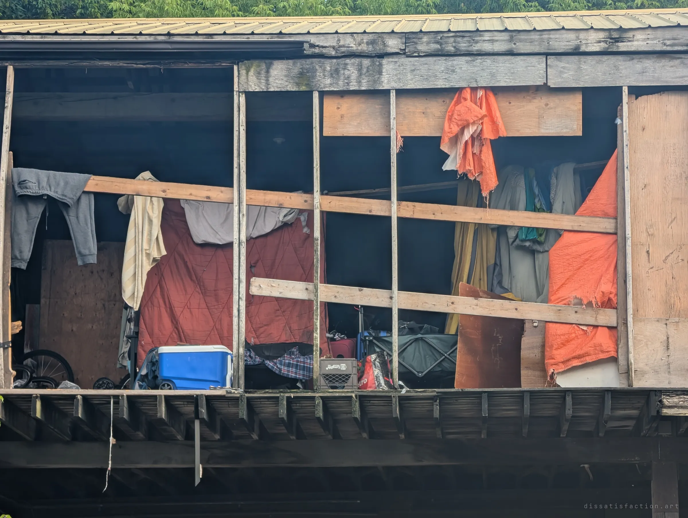 Close view of the shed's upper floor through bare wall studs: a quilt, hanging clothes, torn orange tarps, a blue cooler and a folding cart show a lived-in squat.