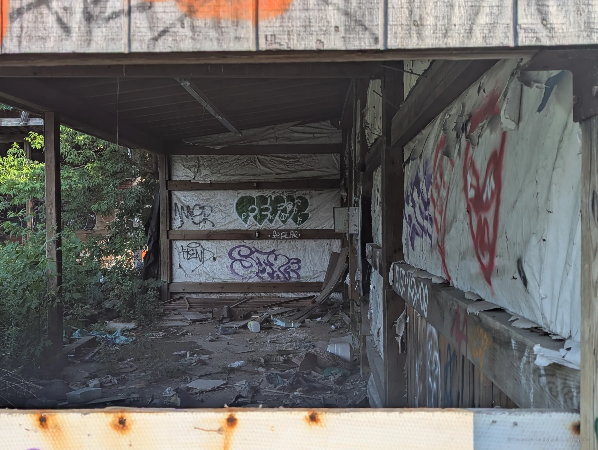 Looking into a covered lumber bay lined with peeling house wrap and graffiti tags, its dirt floor strewn with broken boards and litter.