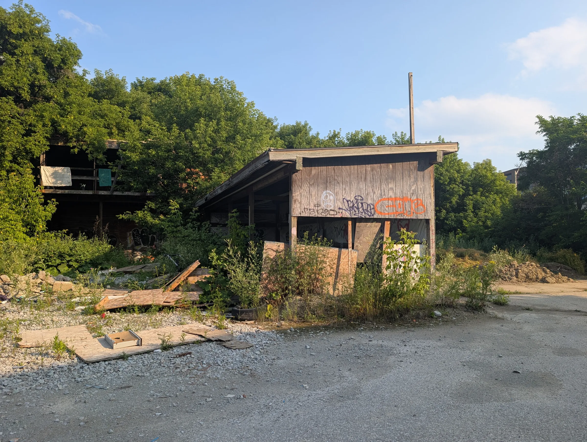 A graffiti-tagged open storage shed and the overgrown two-storey shed behind it, sitting at the edge of a cracked, empty gravel yard.