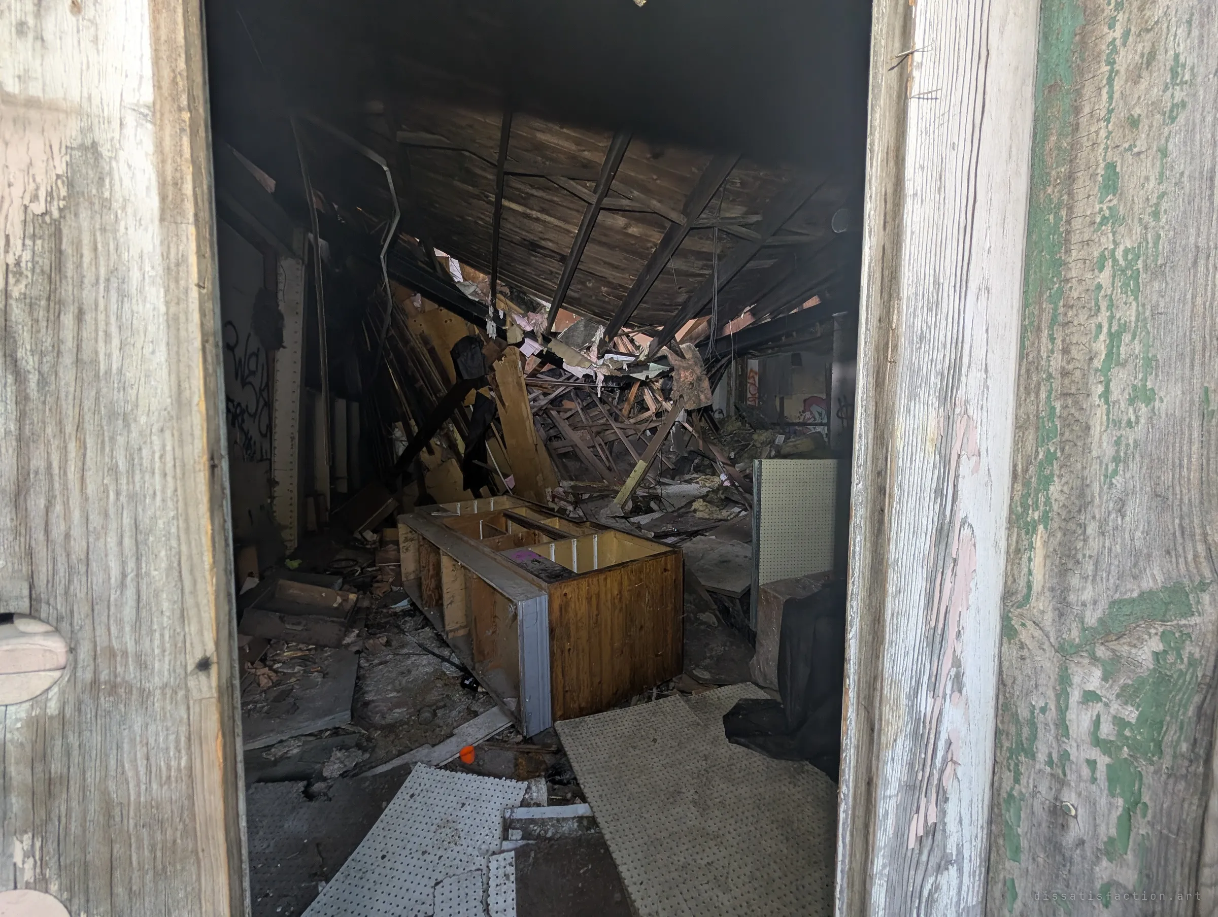 Through a weathered doorway, the inside of the store has caved in: the roof hangs down in splintered timbers over an abandoned wooden counter and pegboard panels.