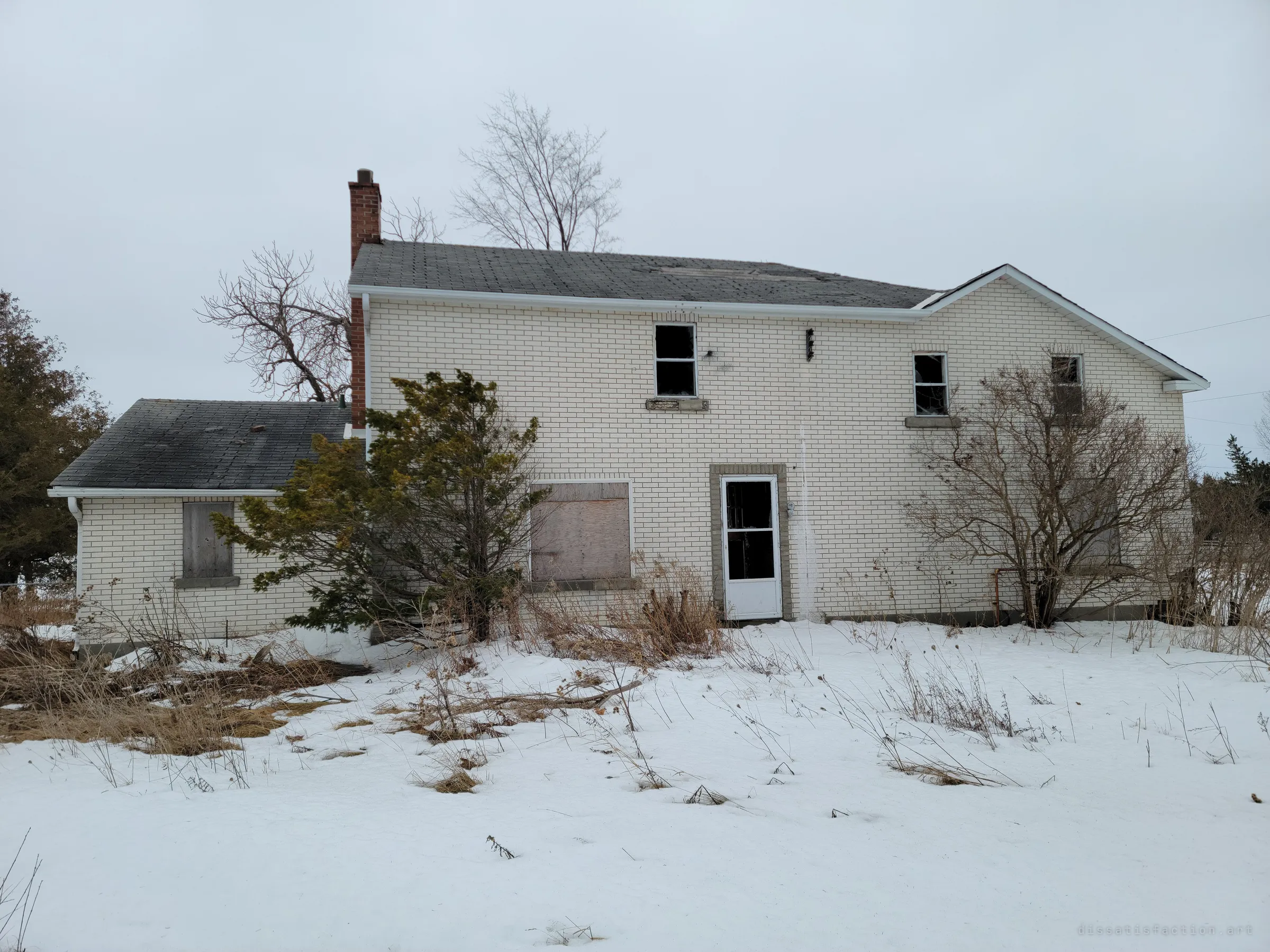 The back of an abandoned two-storey white-brick farmhouse in snow, windows broken or boarded and the yard overgrown. It sets the scene: a family home left to rot through a winter.