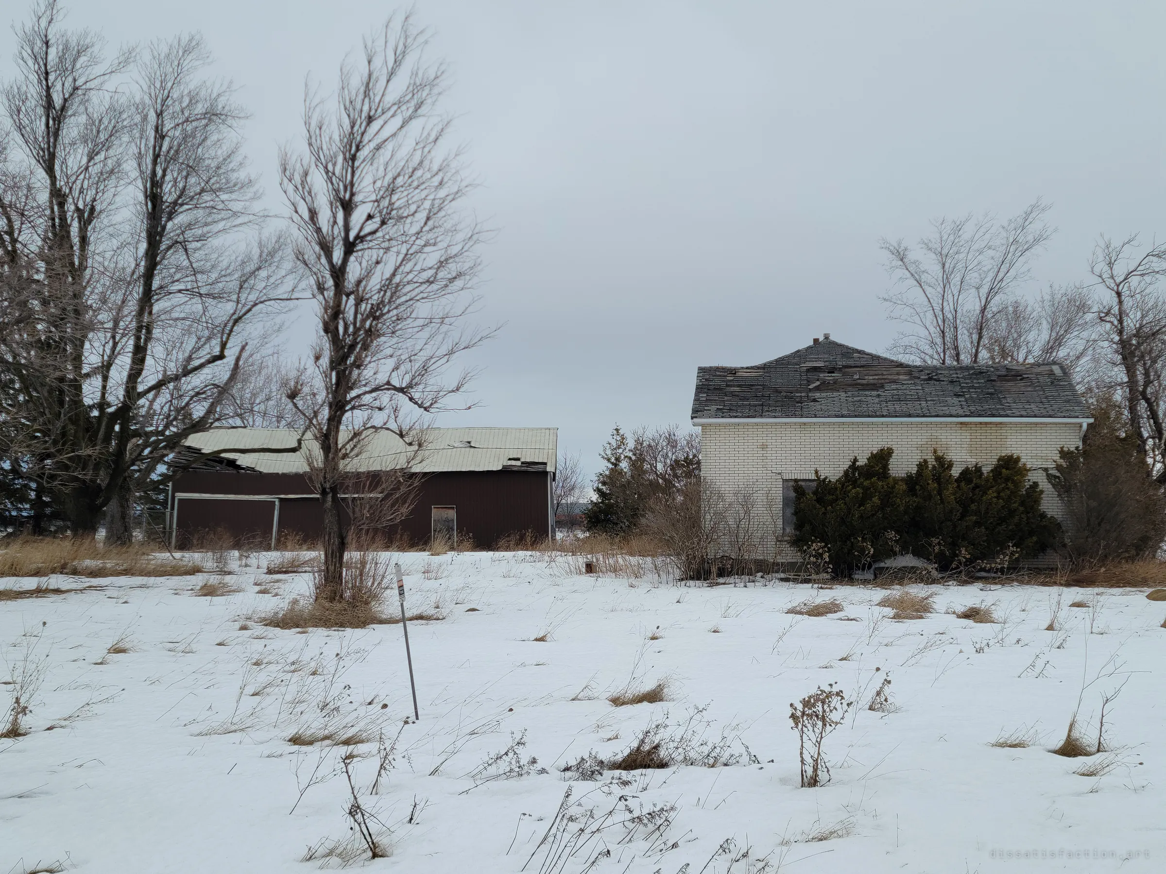 A sagging brown barn and the side of the brick house stand among bare trees across a snowy yard, with a small unreadable utility marker on a post in the foreground. It shows the whole homestead slowly giving way.