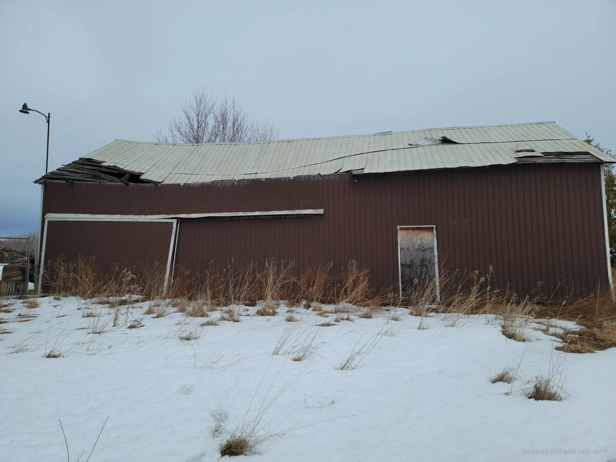 A long brown steel-clad barn whose roof has buckled and caved in at one end, a lone yard light beside it and tall dead grass along its wall. The warped roofline shows how far the structure has failed.