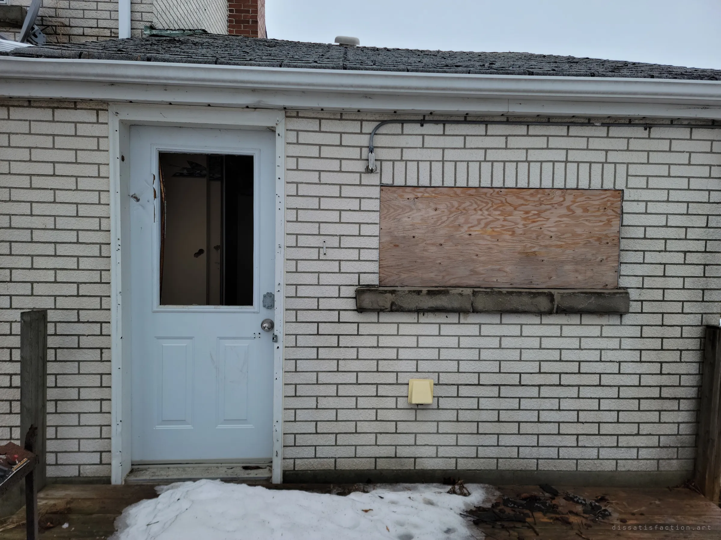 A white side door with a broken window pane next to a boarded-up window on the brick wall, snow drifted on the wooden deck. The smashed glass hints at the break-ins since it was left.