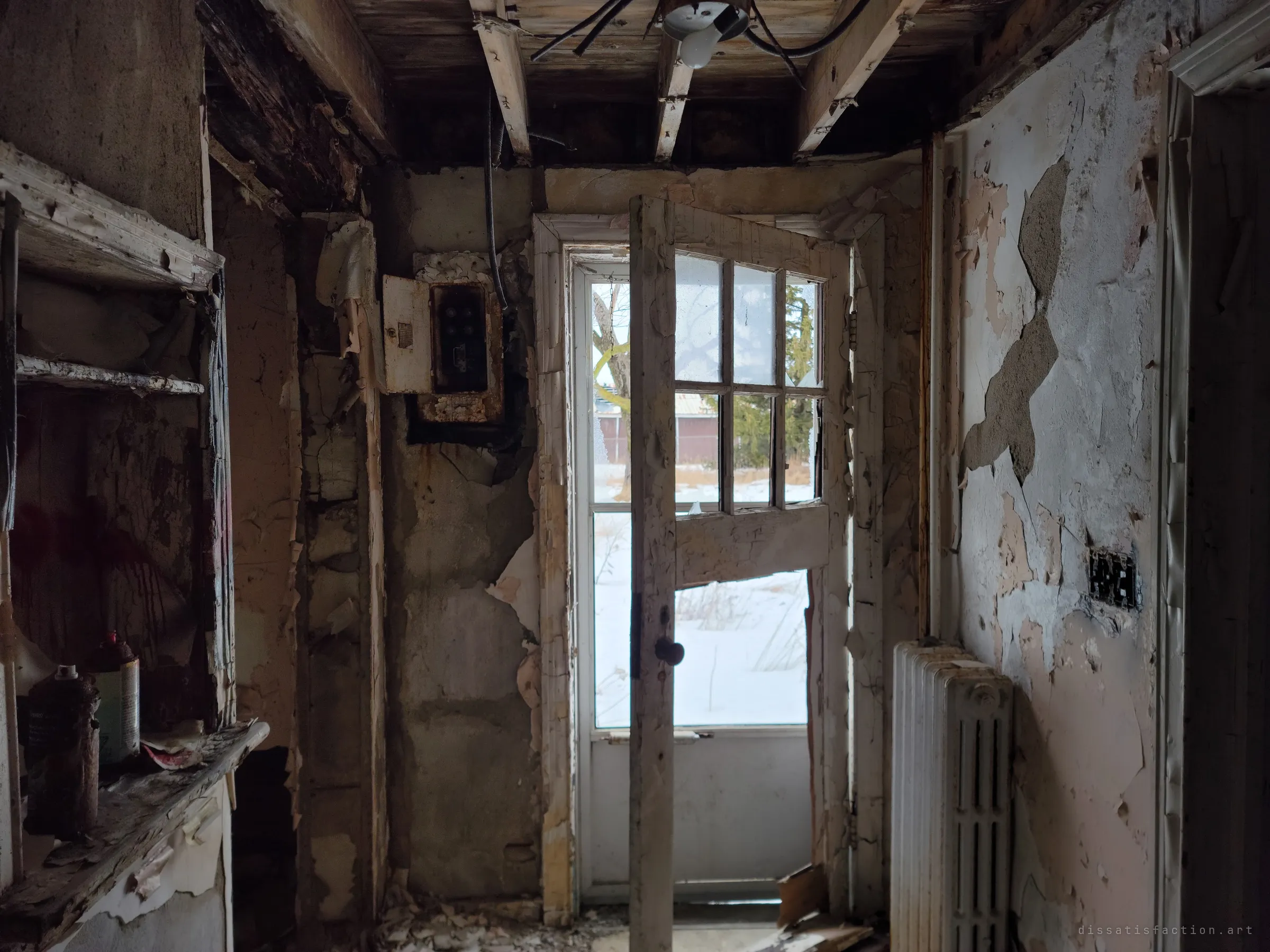 A gutted back entry with peeling paint, an old fuse box and exposed joists, the half-open glazed door letting in snowy daylight. Spray cans on the shelf show the vandals who came before.