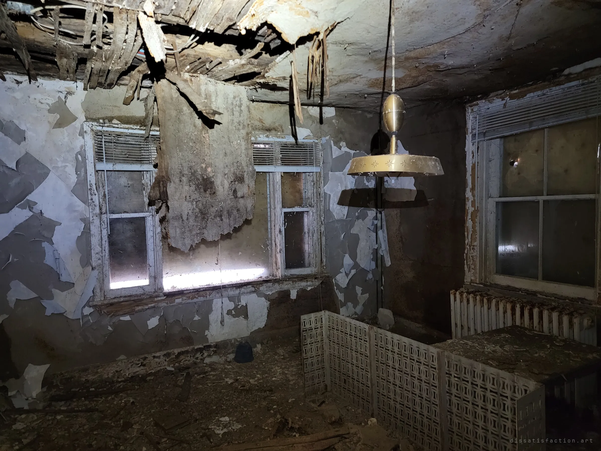 A dark room lit by flash, a brass pendant lamp hanging under a collapsing ceiling, paint peeling off in sheets and a decorative radiator cover by the window. The mid-century lamp is a trace of how the home once looked.