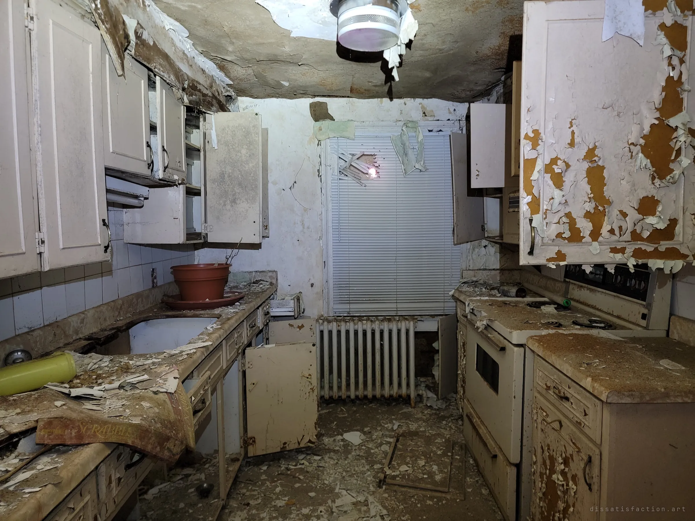 A narrow galley kitchen with cabinet doors hanging open, paint curling off every surface and an old board-game box on the counter. Everyday things left behind make the abandonment feel sudden.