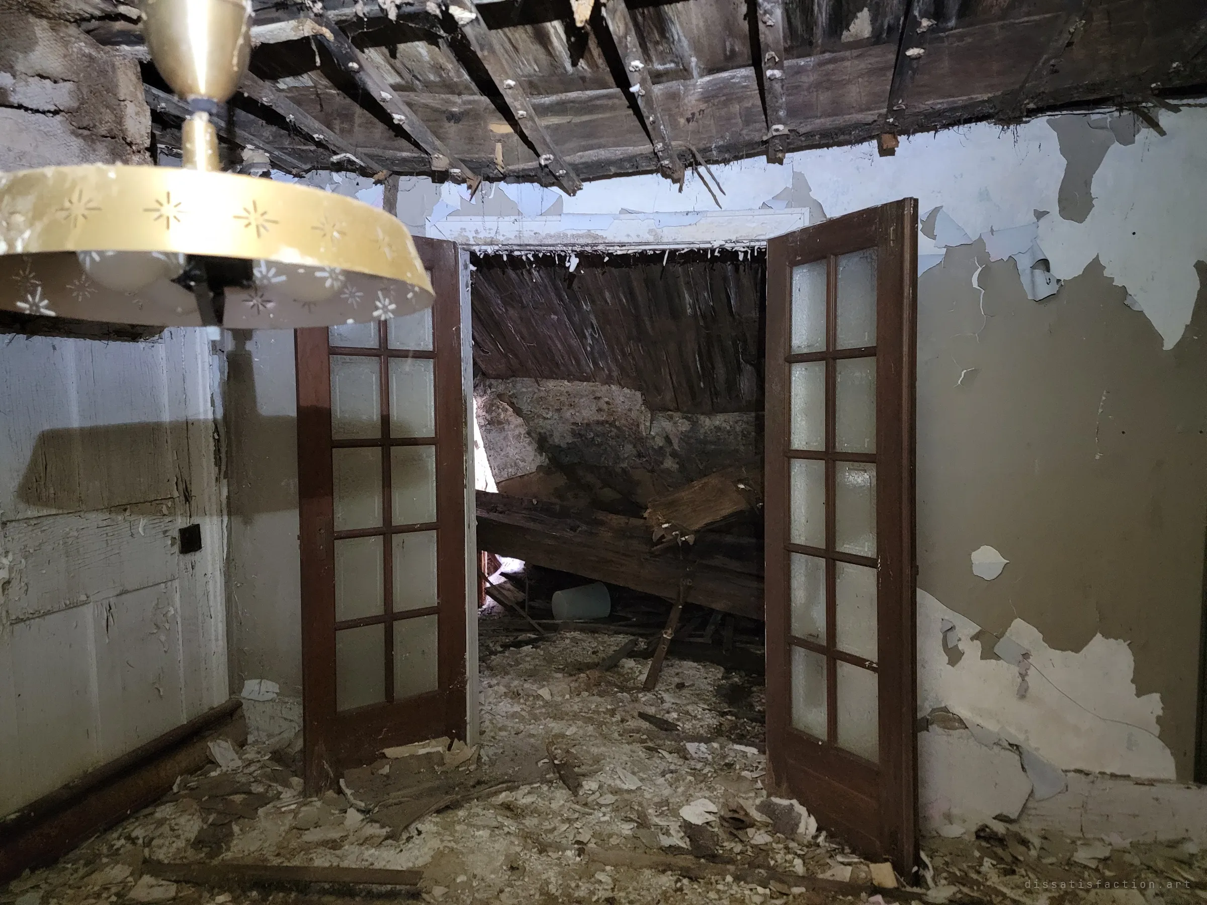 A starburst-patterned ceiling lamp hangs in the foreground while open glass-paned French doors reveal a room where the roof has caved in onto the floor. Stripped ceiling lath and peeling paint show how far the house has fallen apart.