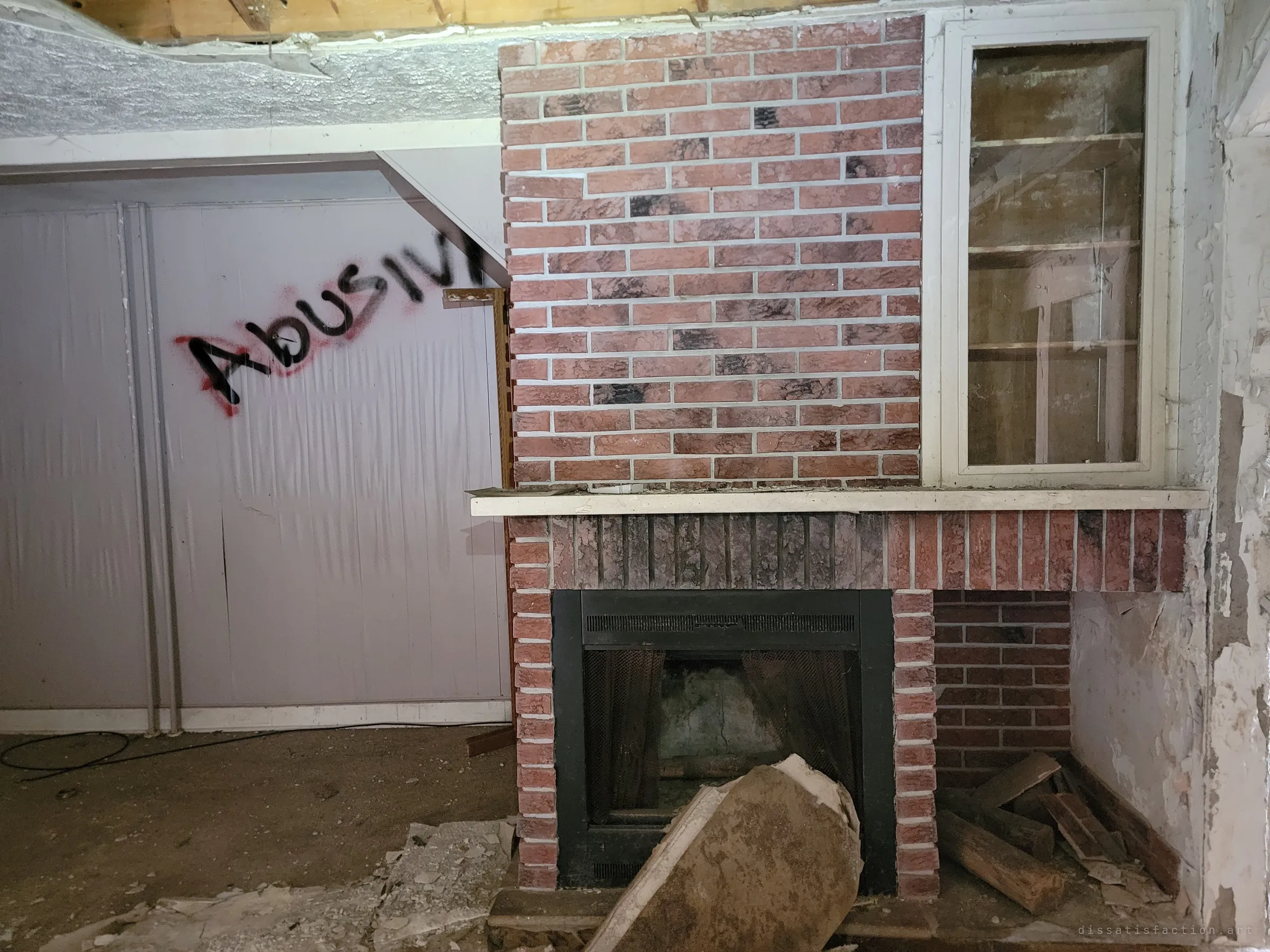 A brick-faced fireplace with an empty glass display cabinet beside it, next to a wall sprayed with the word "abusive" in black and red. Broken plaster and firewood litter the hearth.