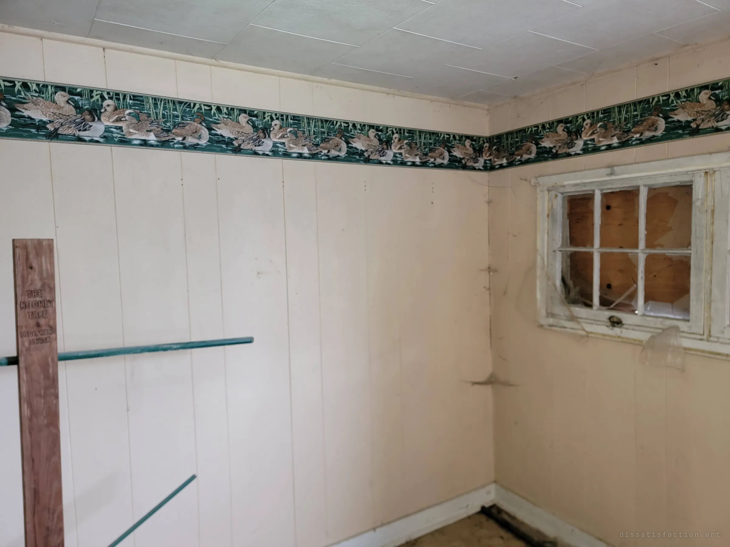 A panelled room with a duck-pattern wallpaper border, a boarded-up window with broken panes and a wooden sports-equipment rack leaning in the corner. The dated domestic details contrast with the decay.