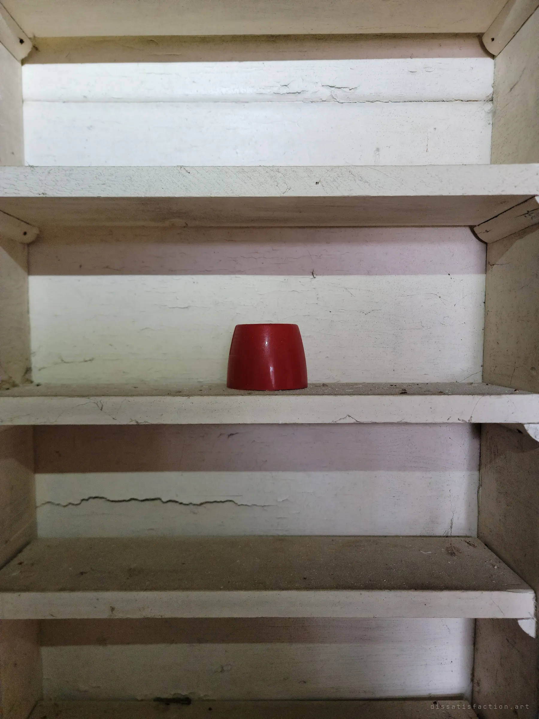 A single red cup-shaped object sits alone on the middle shelf of a dusty white wooden shelving unit. A quiet still life of what was left behind.