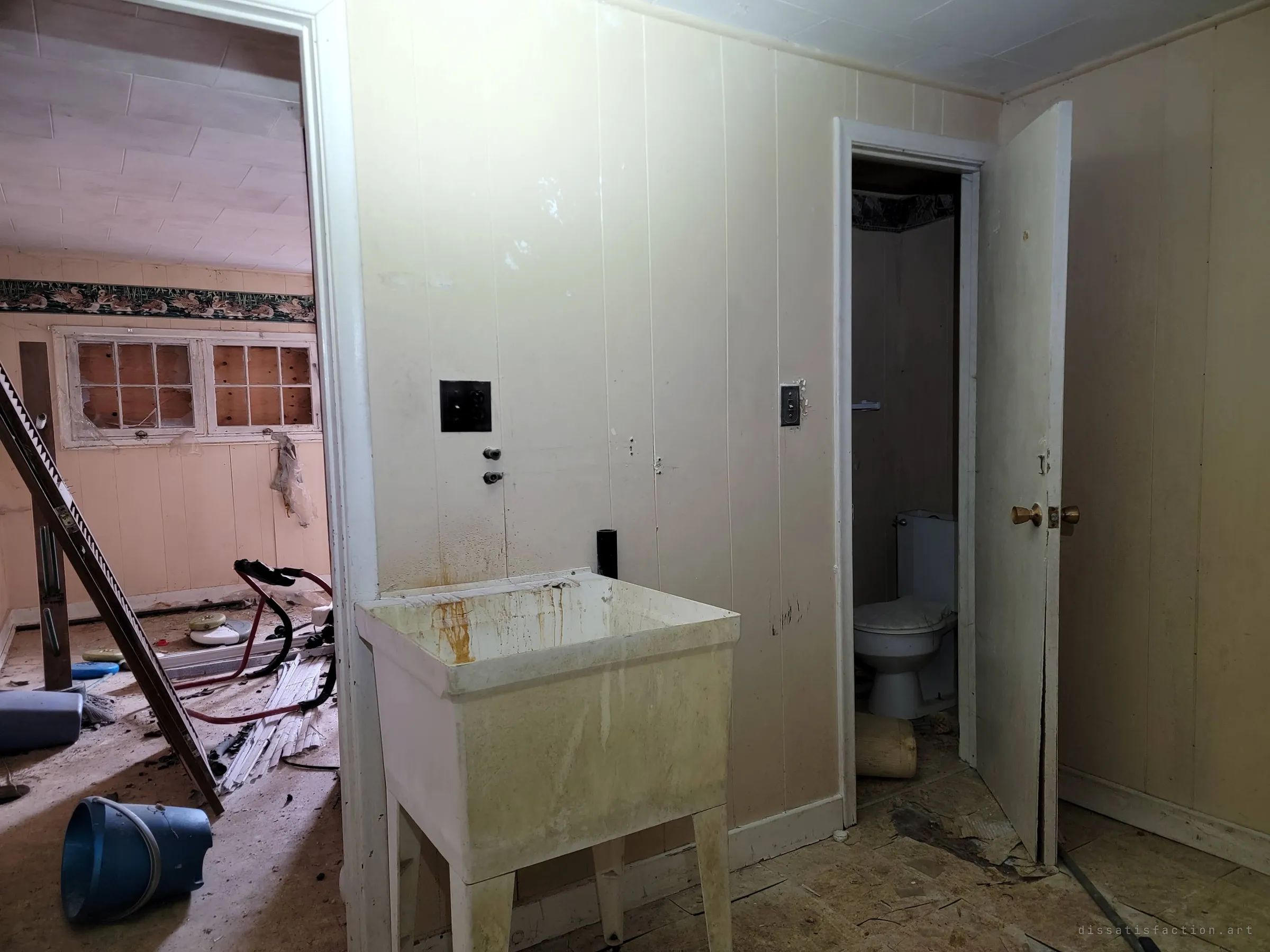 A stained utility sink stands against panelled walls, with a small toilet room through an open door on the right and a debris-strewn room with an exercise machine and boarded windows on the left.