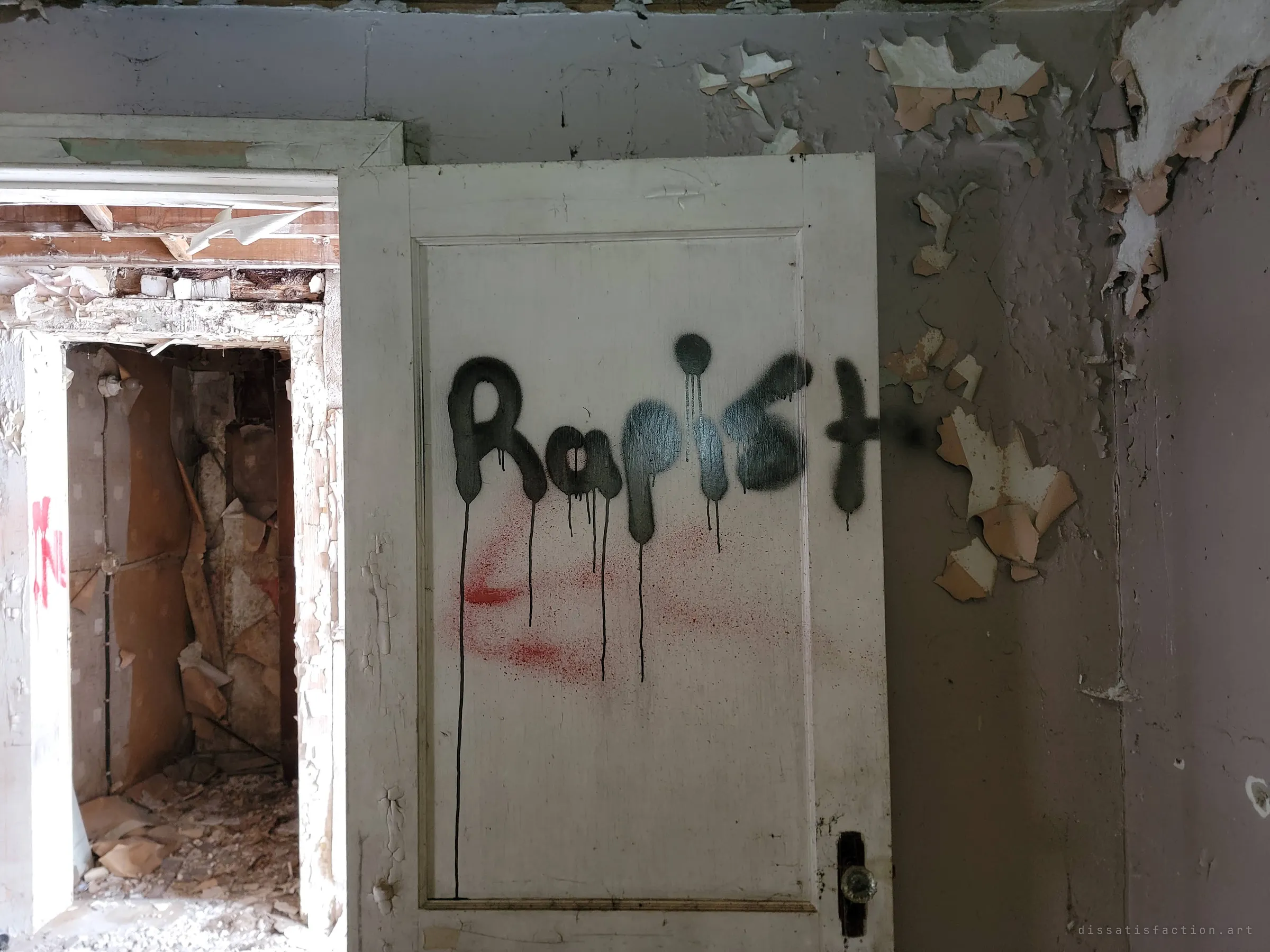A white panelled door sprayed with a black accusatory word in dripping paint, beside a doorway opening onto a gutted room with torn wallpaper. More of the vandals' accusations left on the walls.