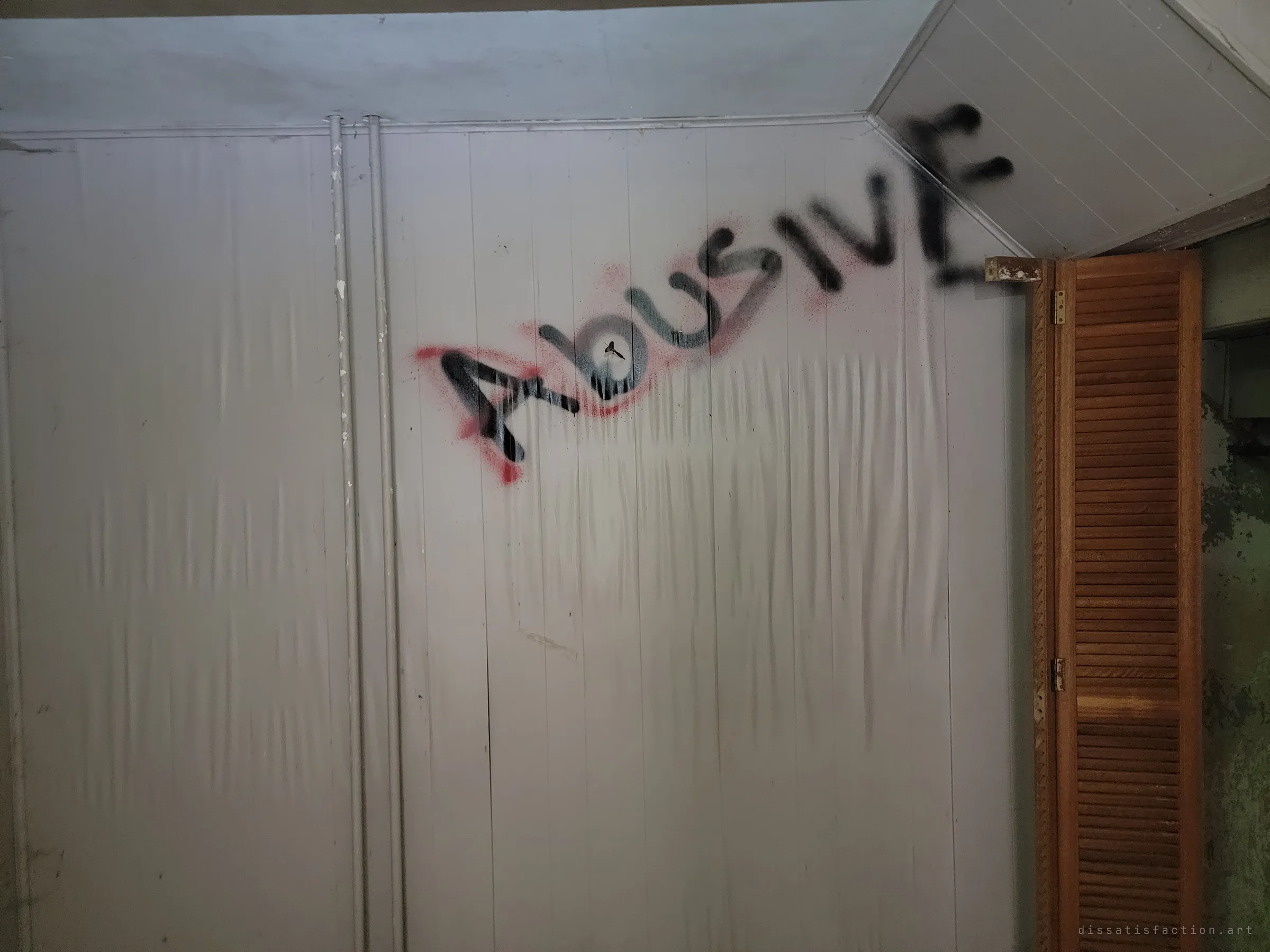 A close view of the plastic-covered wall with "abusive" sprayed in black over red, next to a wooden louvred closet door. The same word seen beside the fireplace, now filling the frame.