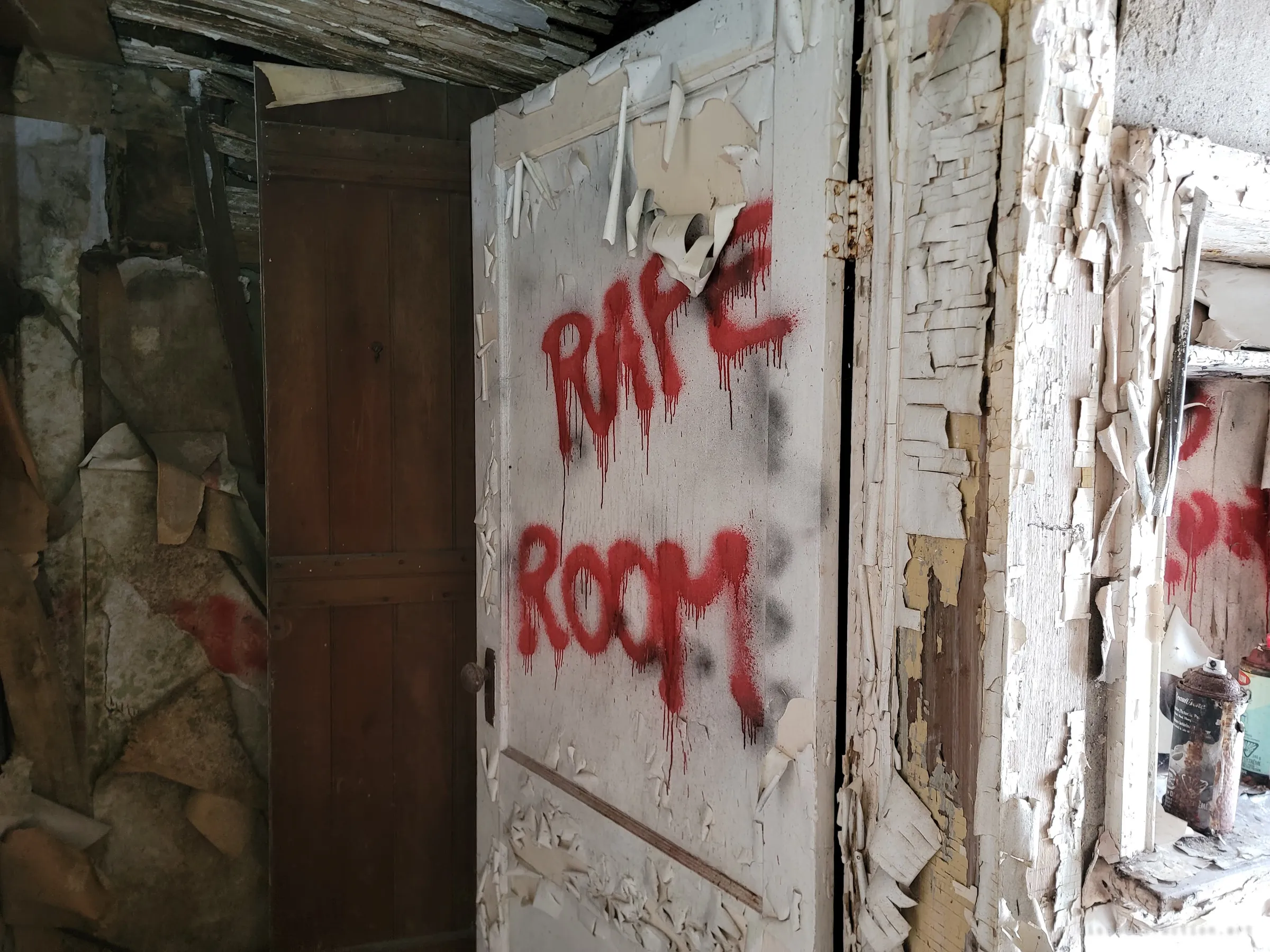 A peeling white door sprayed with dripping red letters labelling the room, with abandoned spray-paint cans sitting on a window ledge to the right. The cans suggest the graffiti was done on site by visitors.