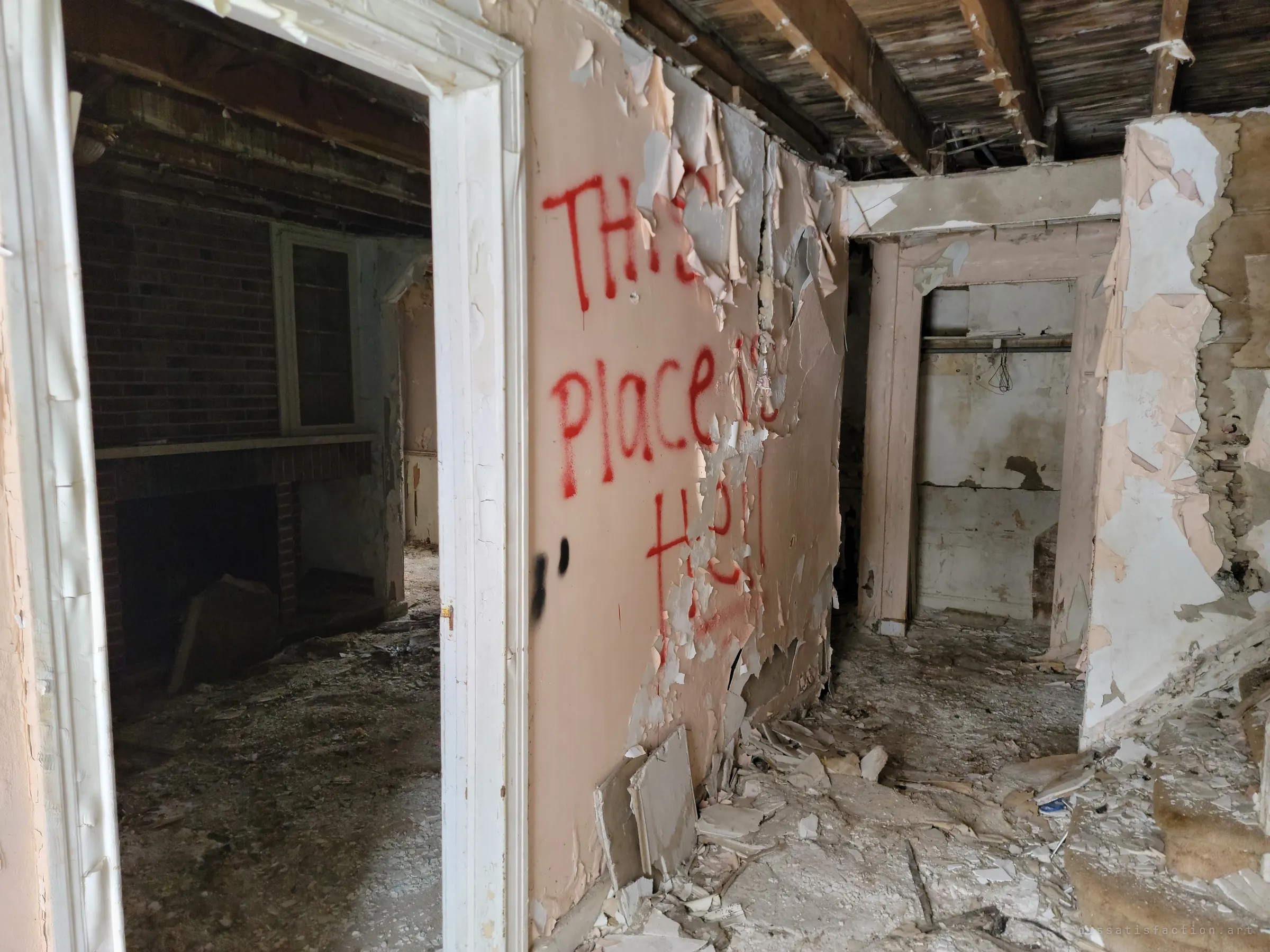 A gutted hallway with peeling pink paint, collapsed ceiling boards and red spray-painted graffiti declaring the place is hell, beside a doorway into a brick-walled room with a fireplace.