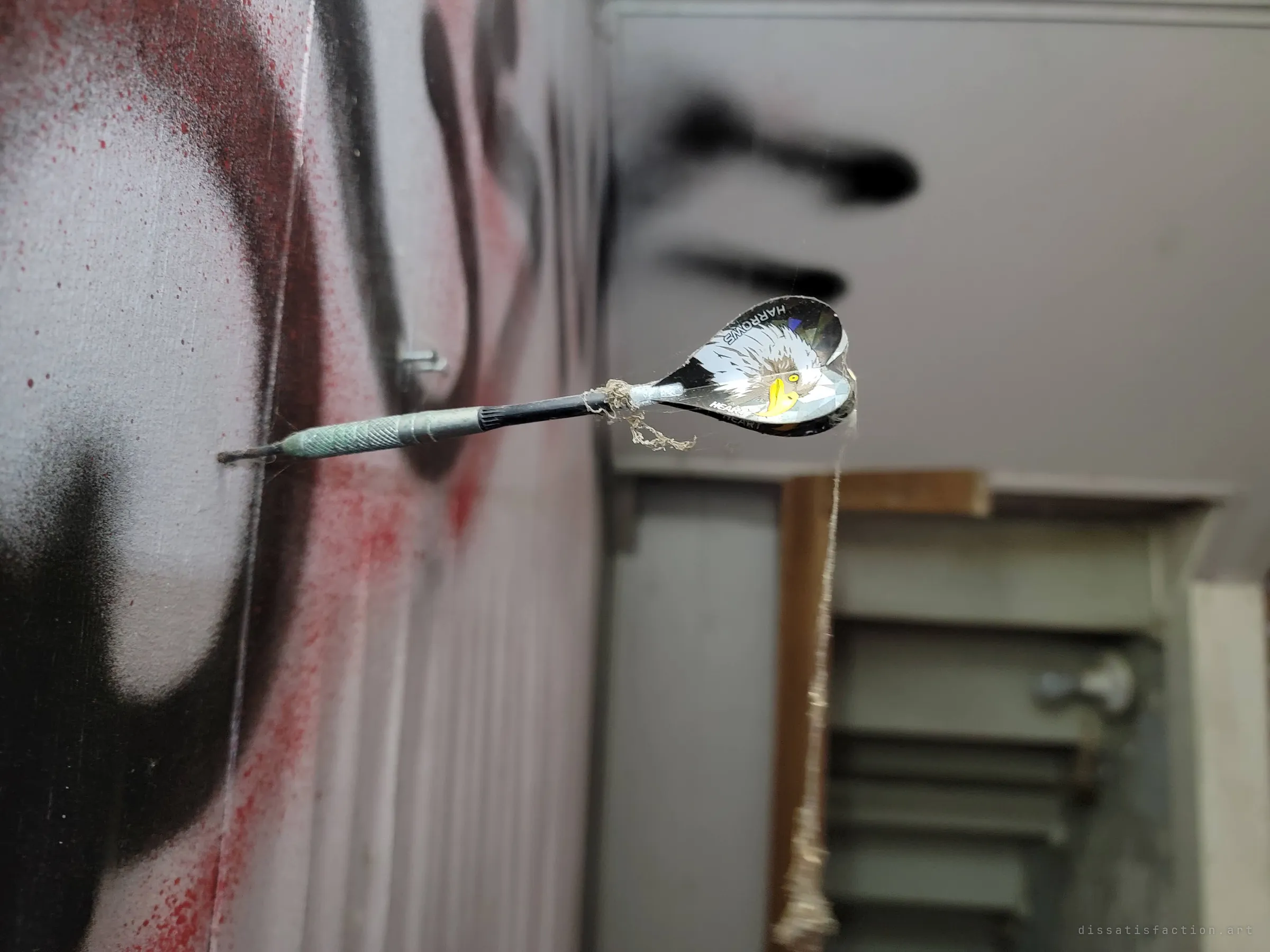 Close-up of an old dart stuck in a graffiti-covered wall, its eagle-printed flight wrapped in cobwebs, with a blurred doorway behind it.