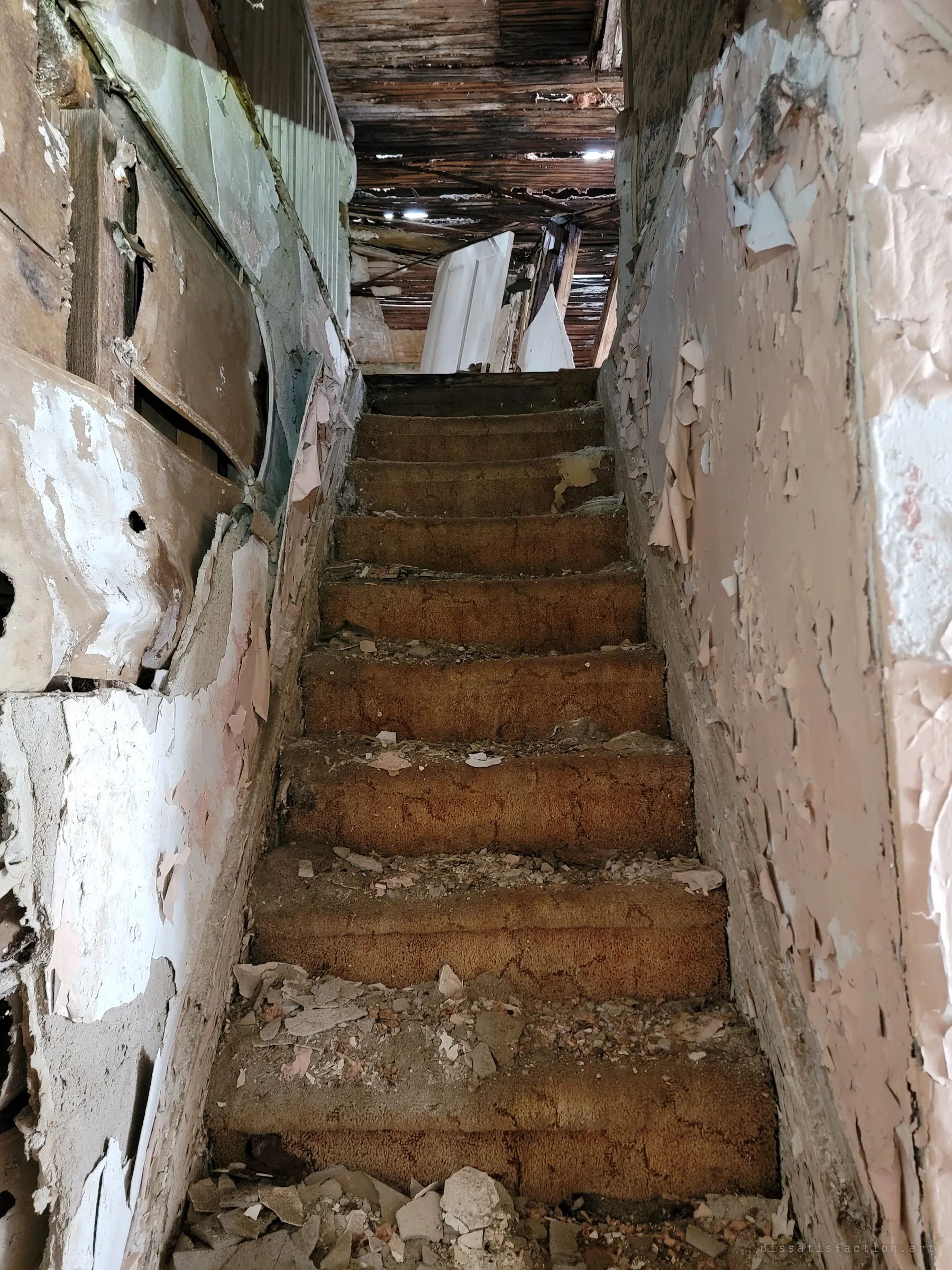 Looking up a carpeted staircase littered with fallen plaster, walls shedding paint on both sides and a broken ceiling and leaning doors at the top.