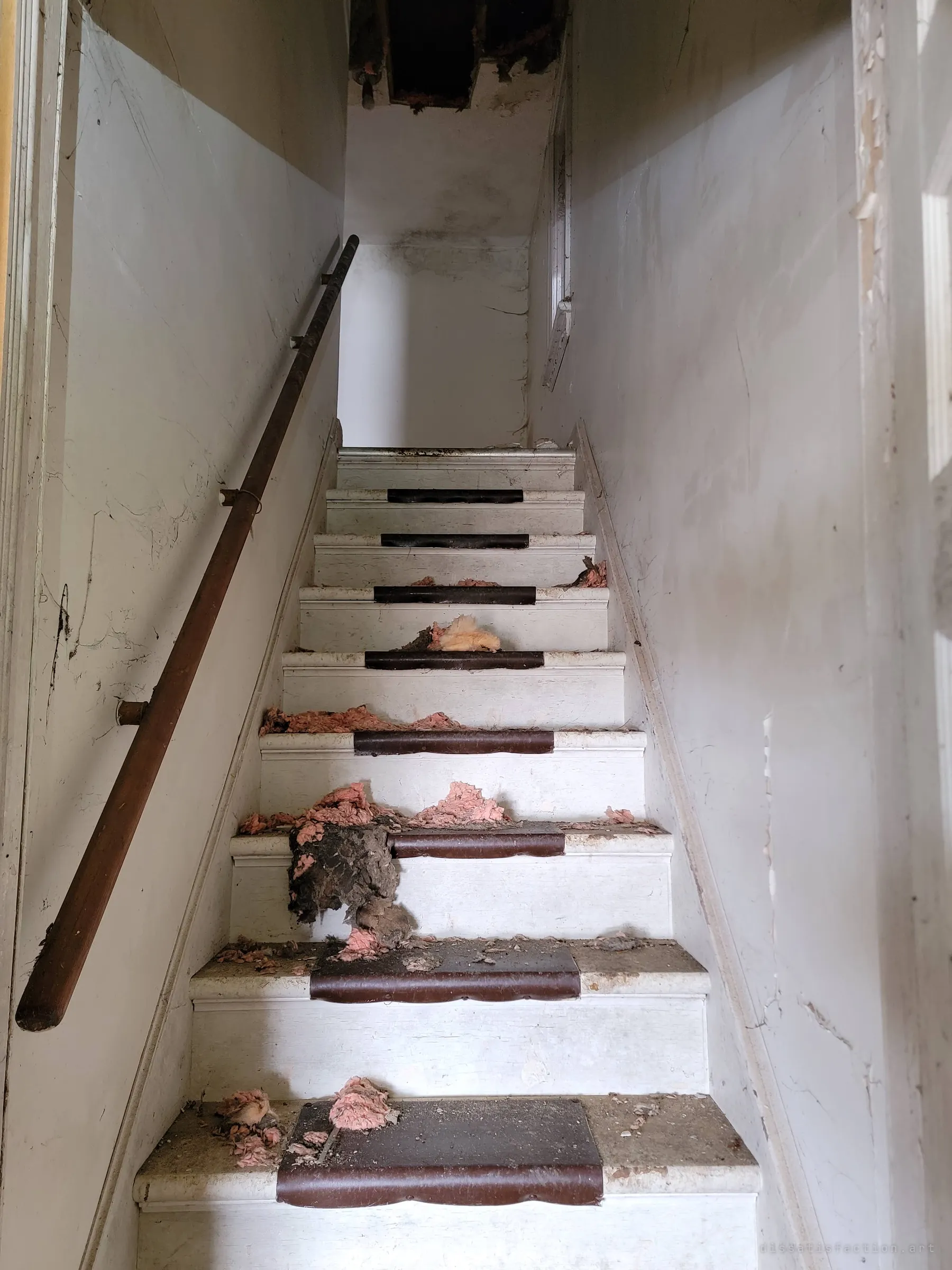 A narrow white stairwell with worn brown tread covers and clumps of pink insulation scattered down the steps from a hole in the ceiling above.
