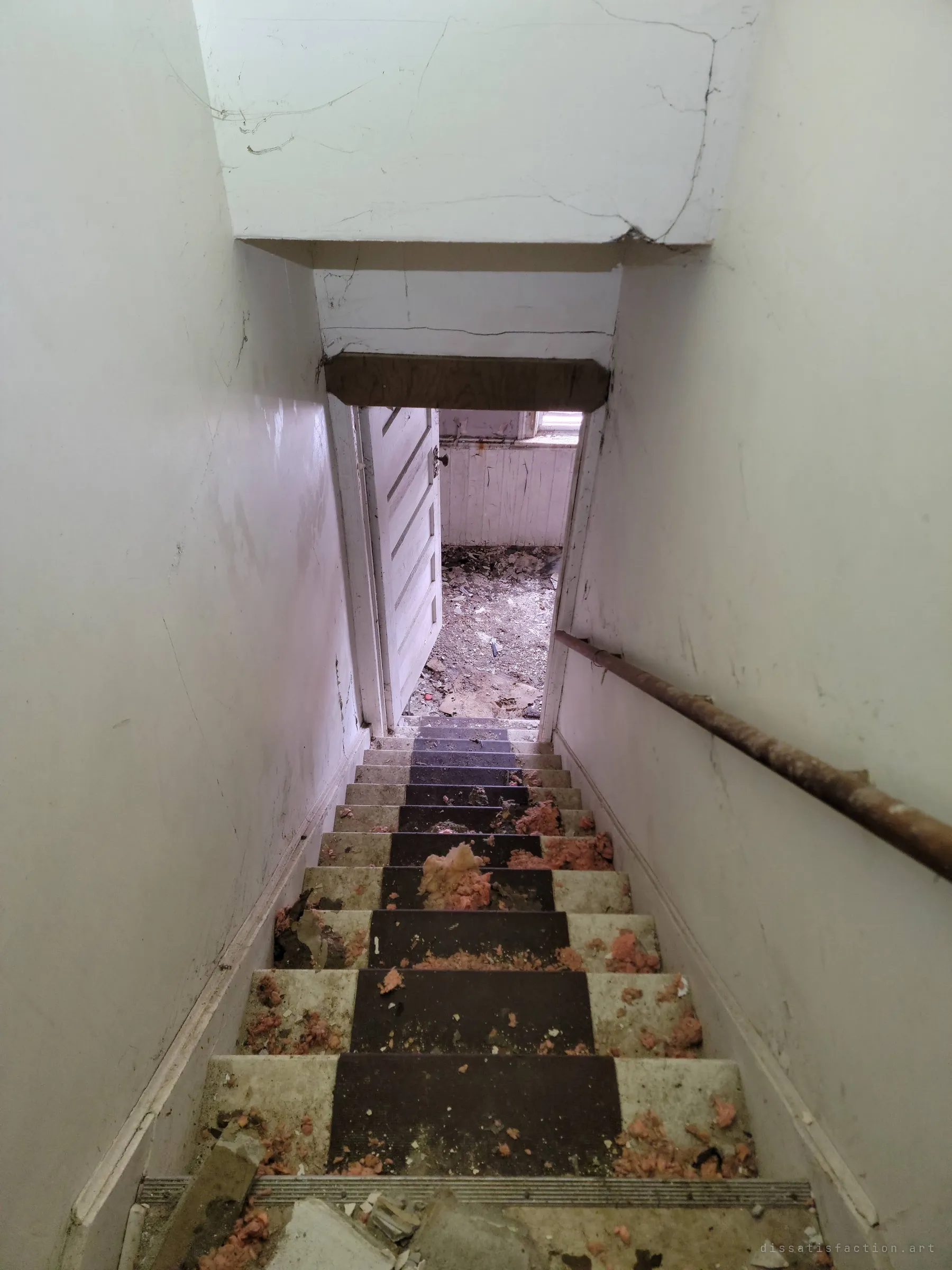Looking down the same cramped stairwell toward an open door and a debris-strewn room below, insulation scattered over the steps.