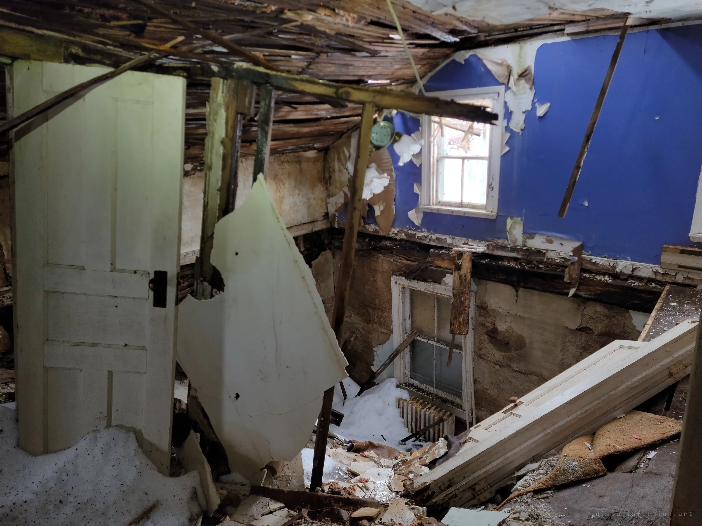 A room with a caved-in floor and ceiling: a bright blue wall above, snow drifted inside, doors and beams collapsed into the basement below.