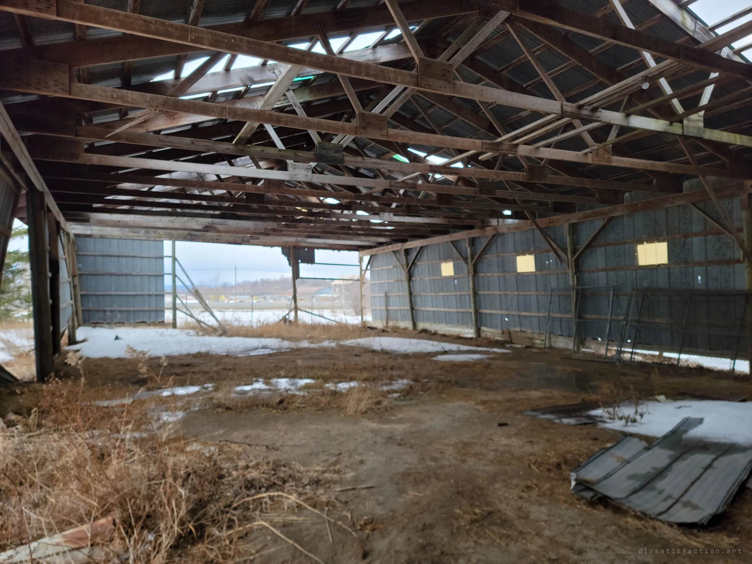 Inside an open-sided pole barn with a sagging truss roof and missing panels, snow patches on the dirt floor and a pale winter landscape beyond the open end.