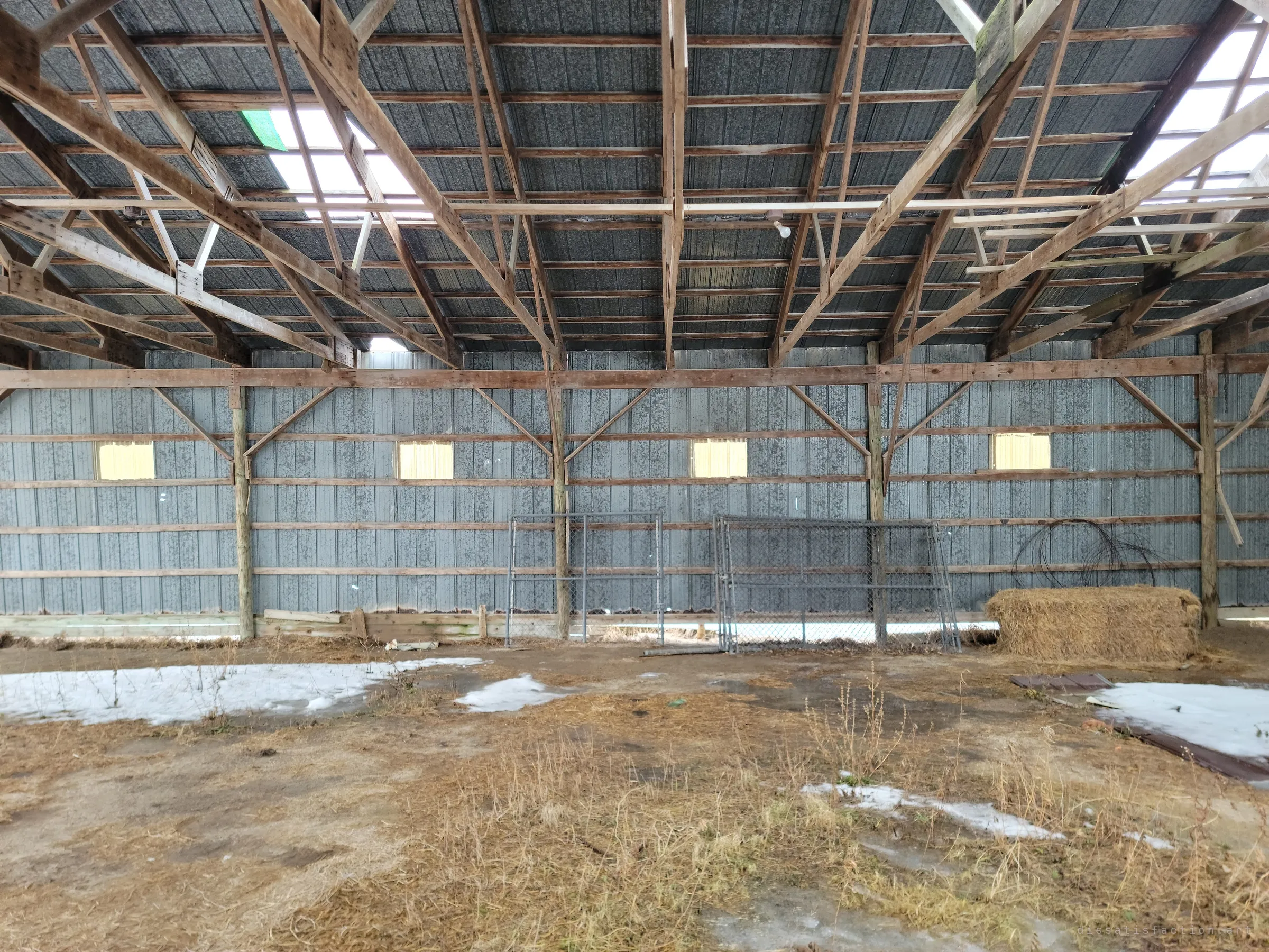 The long metal wall of the same barn, lit by small yellow windows, with chain-link panels, a hay bale and coiled wire beneath the timber trusses.