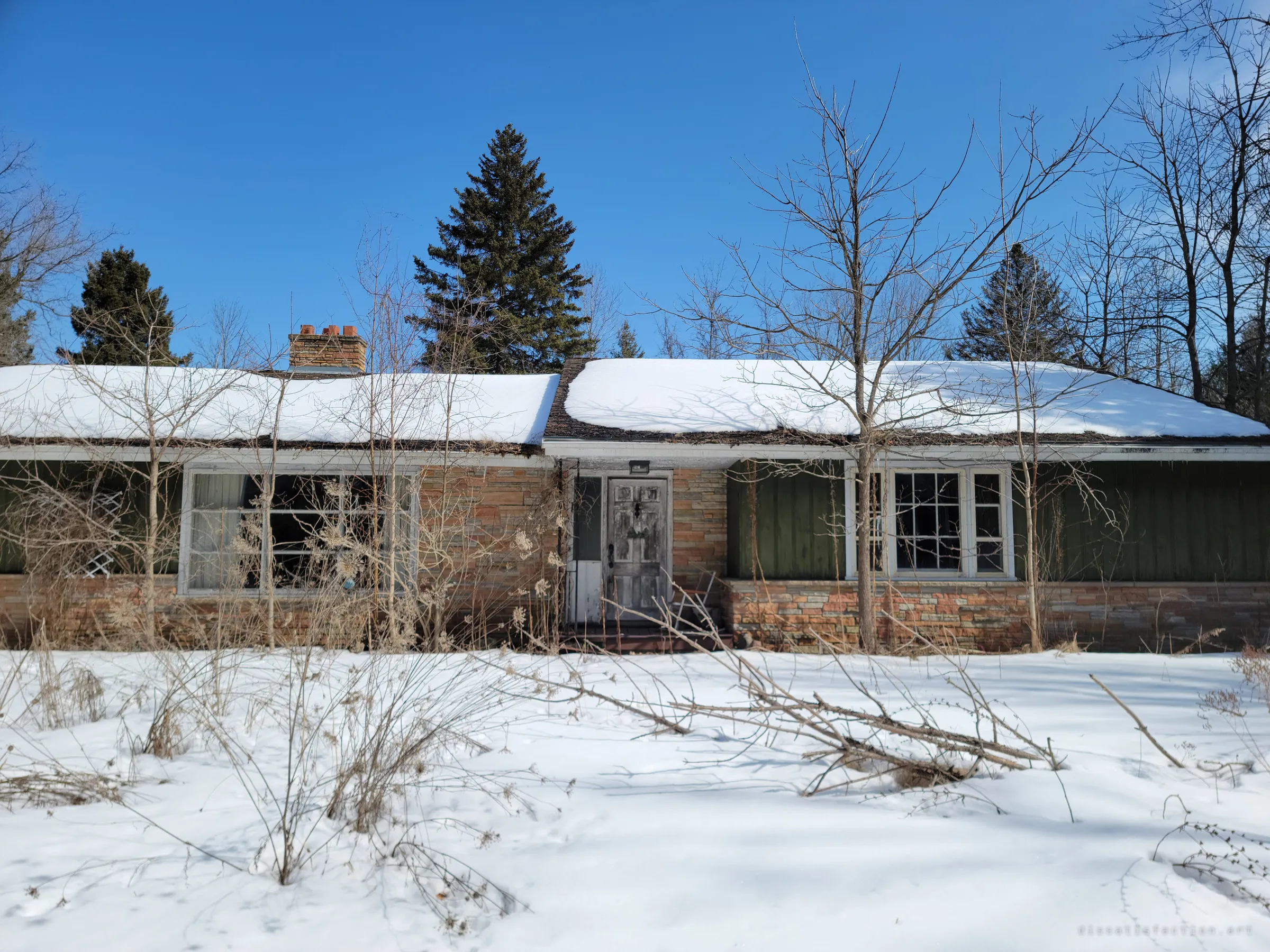 The snowed-in front of a low mid-century bungalow, stone and green siding half hidden behind bare saplings. The weathered front door and overgrown yard show how long it has been left.