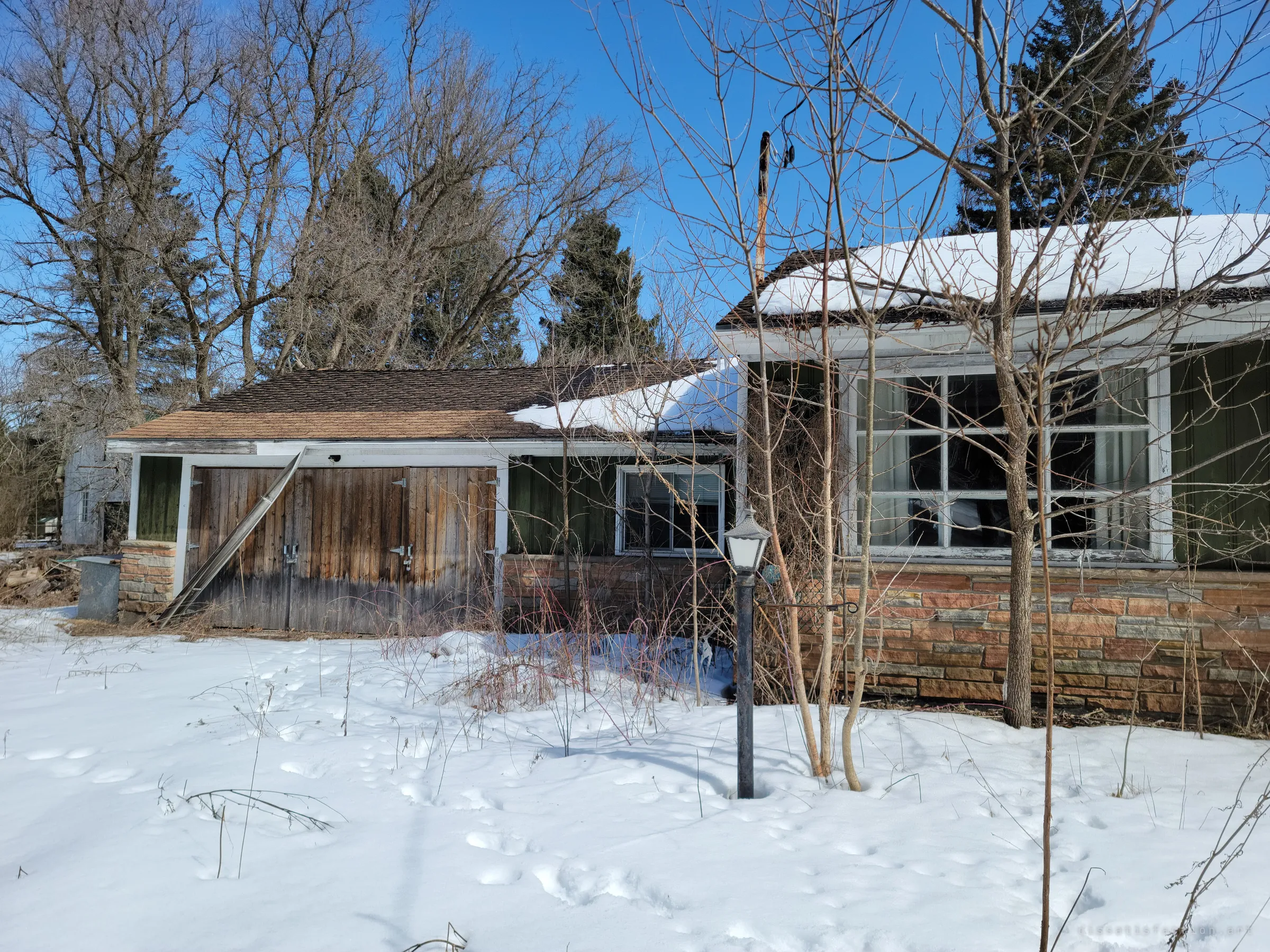 An attached garage with weathered plank doors beside the main house, a lone lamp post standing in deep snow. A ladder leans against the doors and saplings crowd the windows.