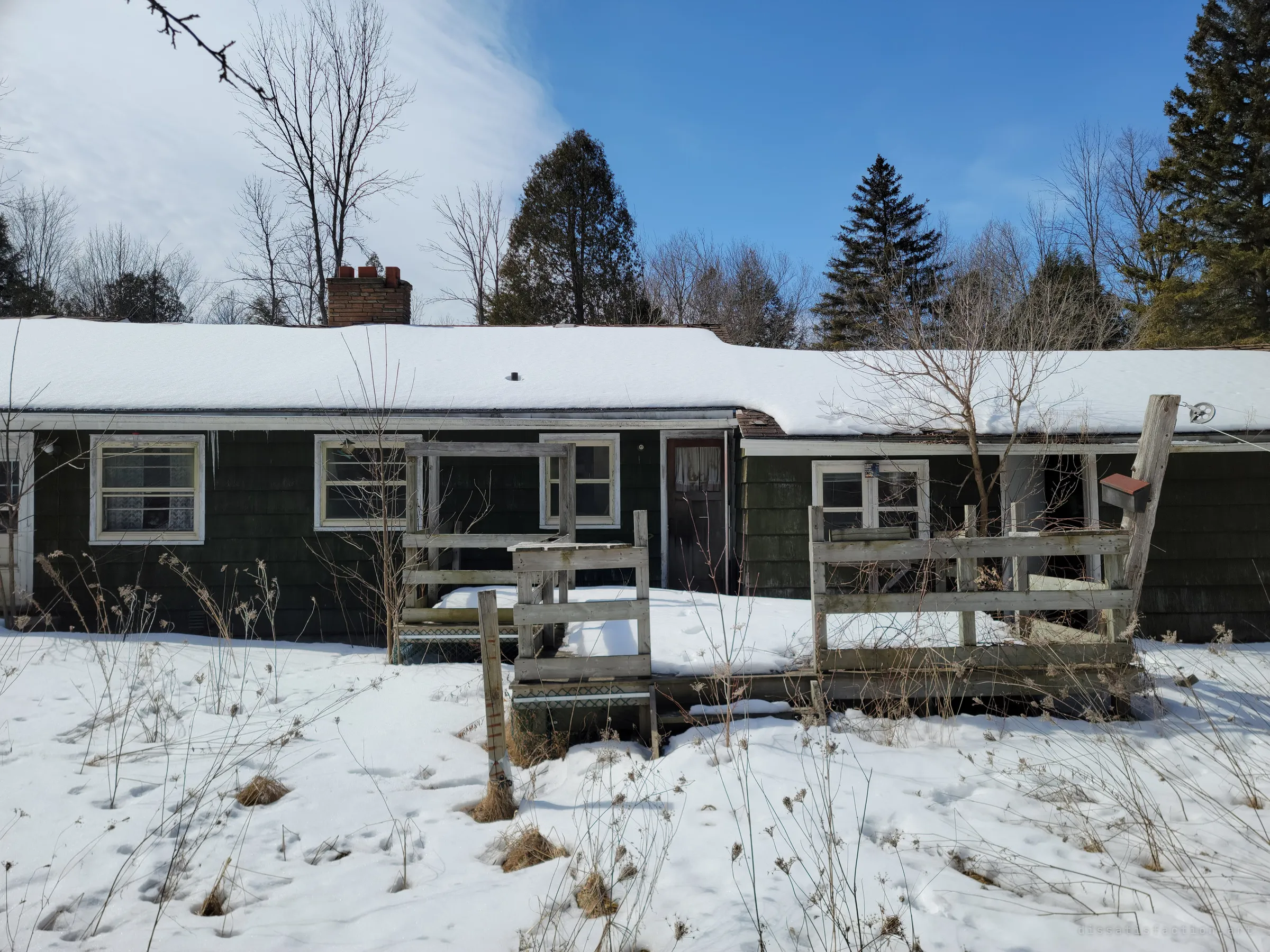 The back of the house with dark shingle siding and a collapsing wooden deck half buried in snow. A small mailbox hangs on a rough post beside a clothesline pulley.