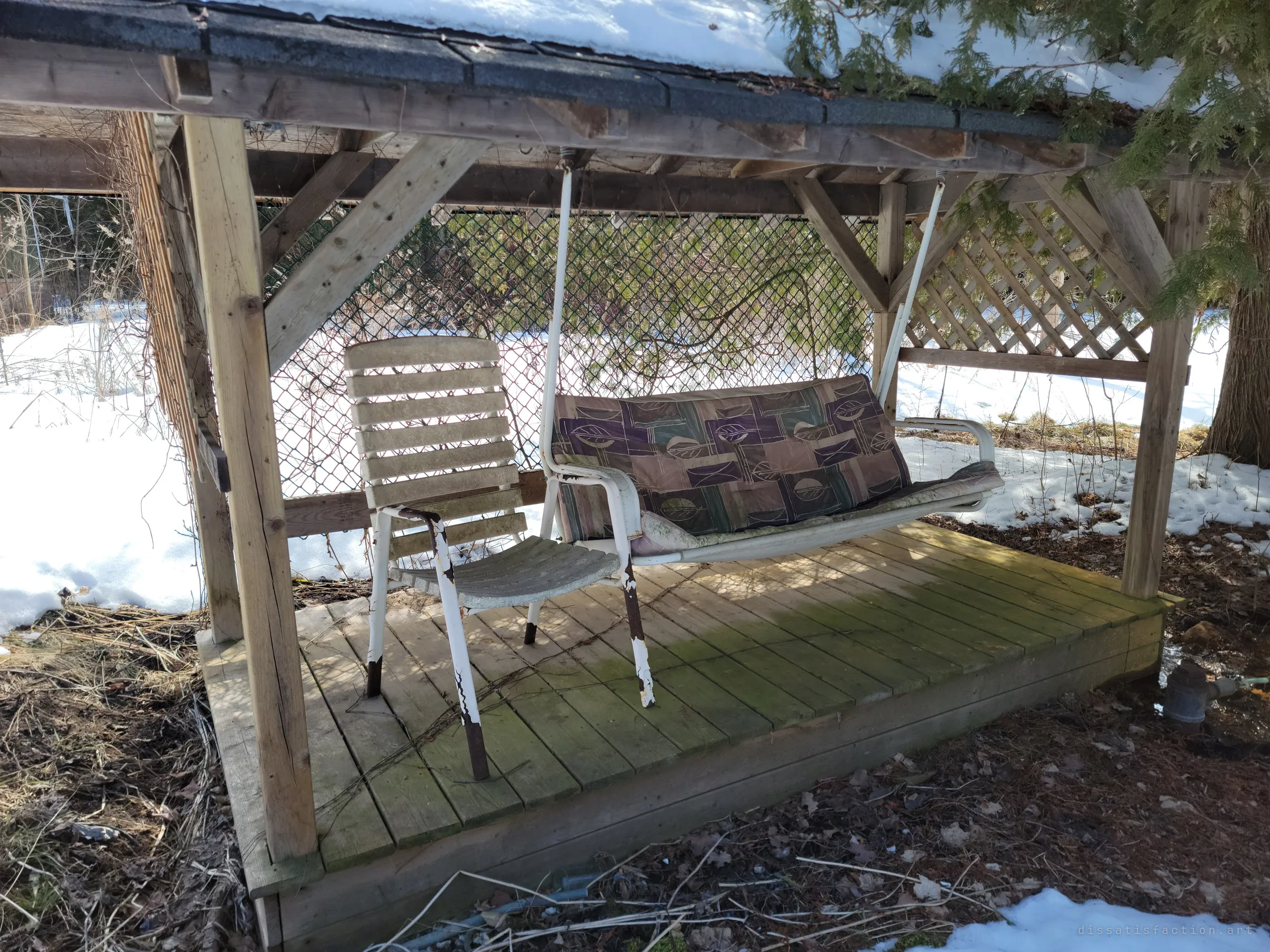A roofed porch swing with faded patterned cushions and a peeling slatted chair on a mossy wooden platform. The snow around it has started to melt back.