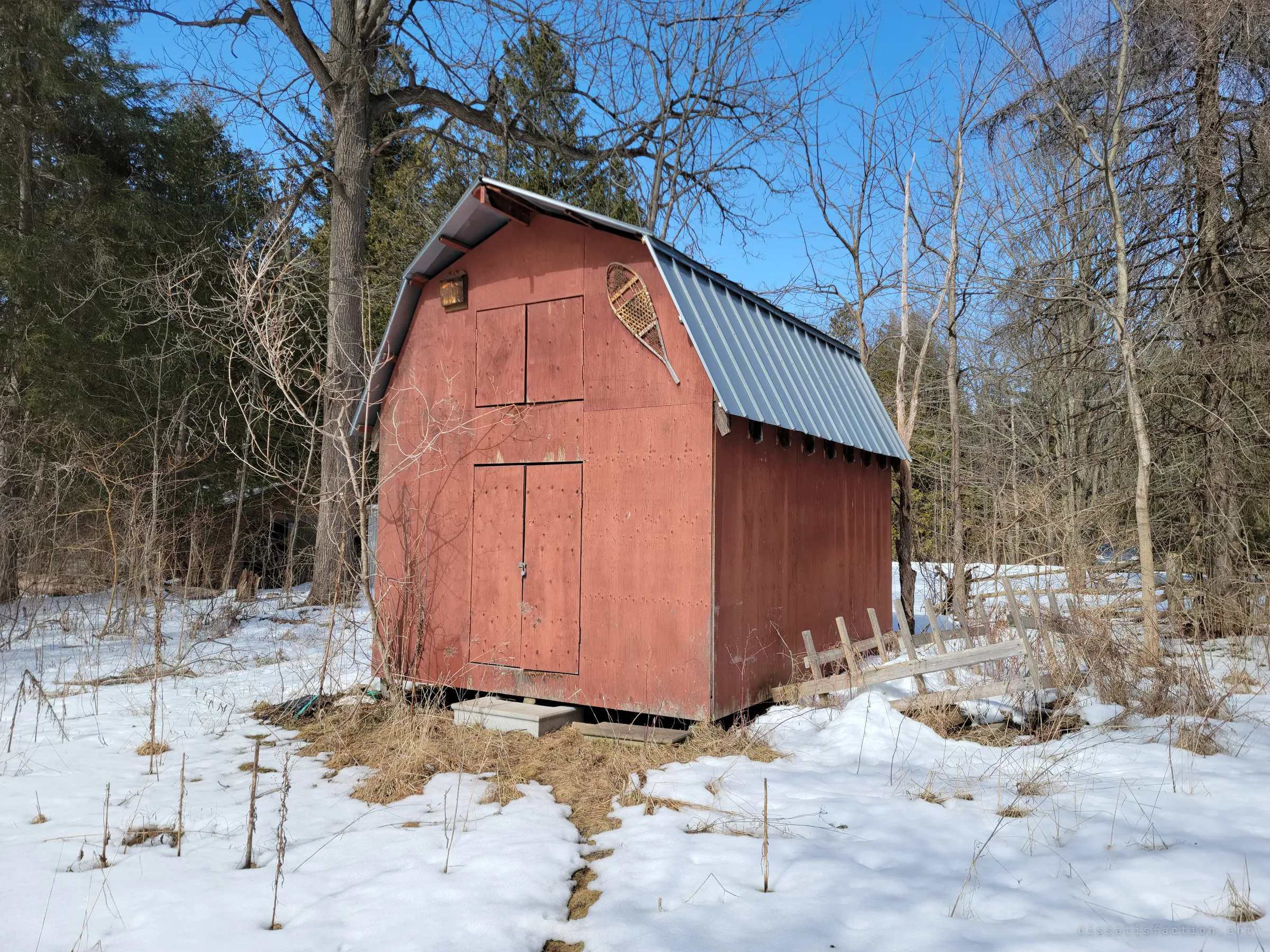 A small red barn-style shed with a metal roof at the edge of the woods, an old snowshoe hung on its gable. A trampled path in the snow leads up to its doors.