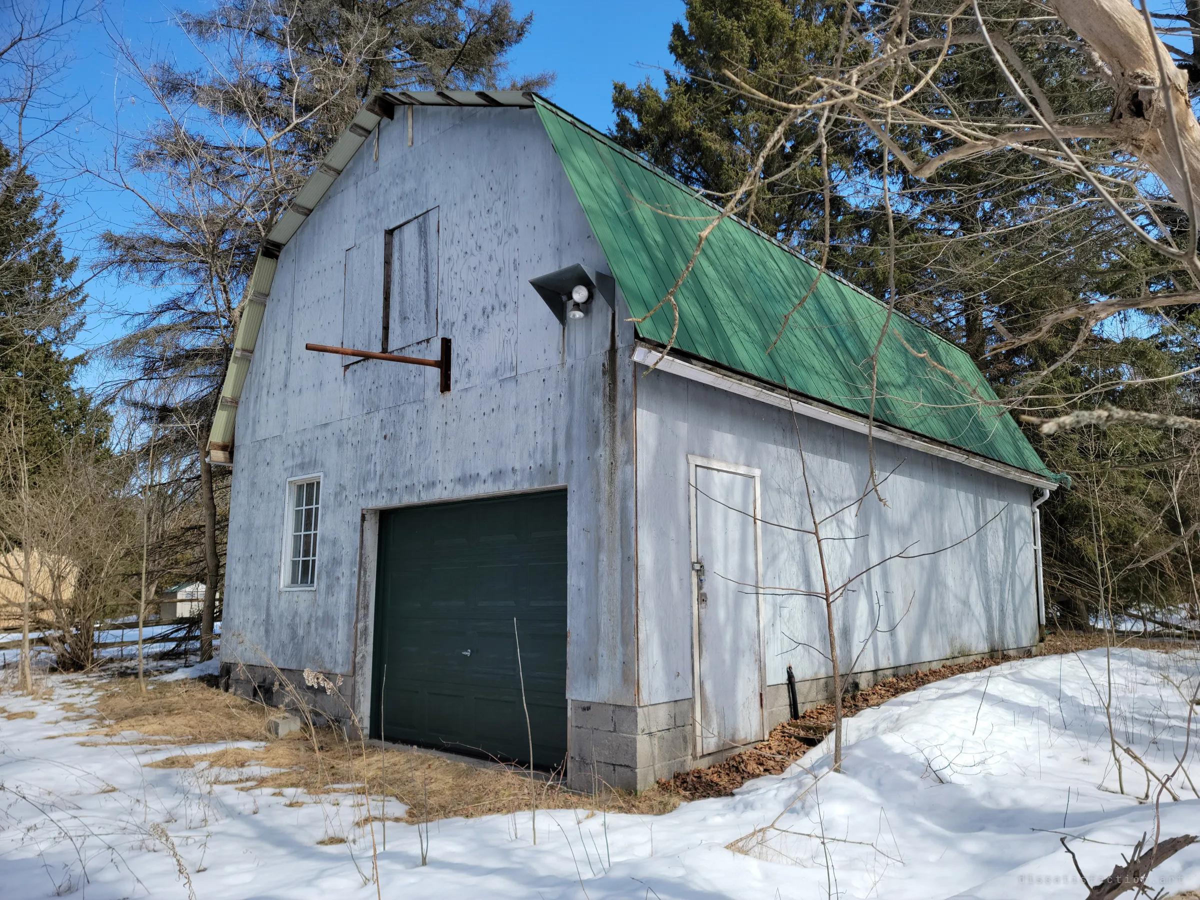 A tall grey gambrel-roofed garage with a green metal roof and a dark garage door, tucked among evergreens. A rusted hoist arm still juts from the loft door.