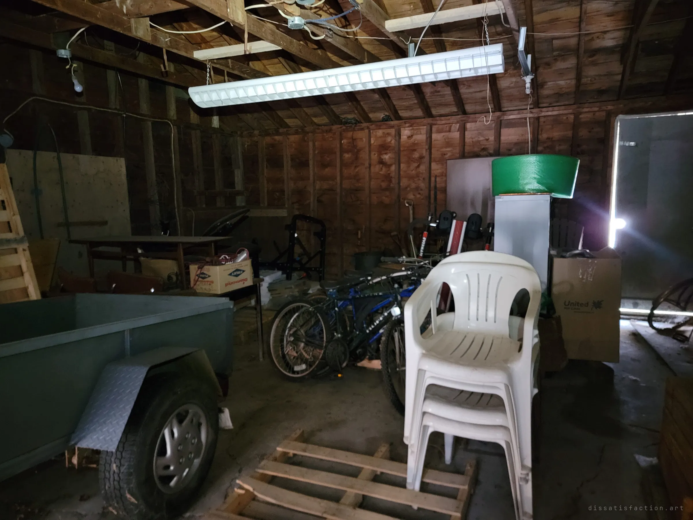 Inside the garage: a utility trailer, stacked plastic lawn chairs, bicycles and boxes under a hanging fluorescent light. Daylight spills in through the open side door.
