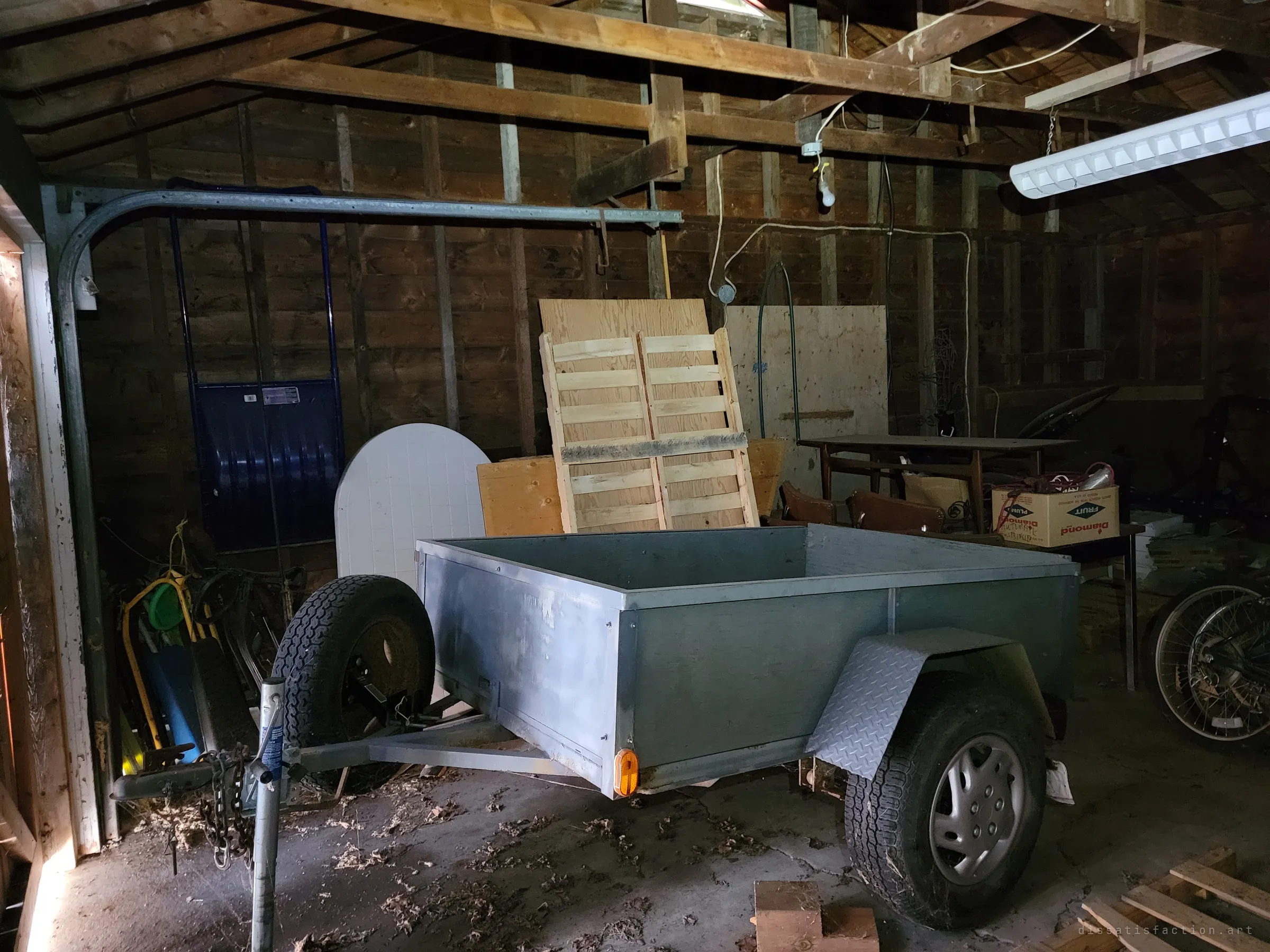 A galvanized utility trailer parked in the dim garage, surrounded by pallets, a round table top and old sleds. The garage door track curves overhead.