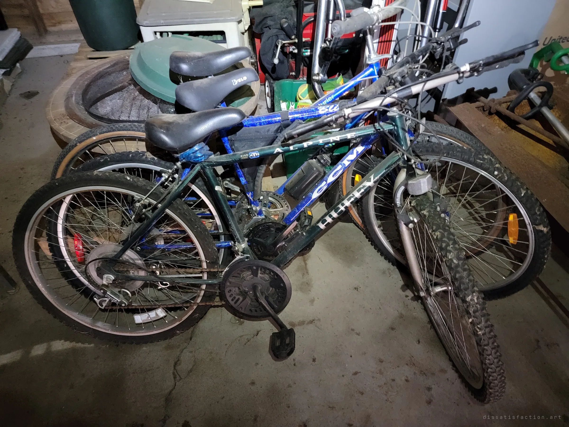 A tangle of old mountain bikes leaning together on the garage floor, lit by a flashlight. Their muddy tyres and taped frames suggest years of use before they were left behind.