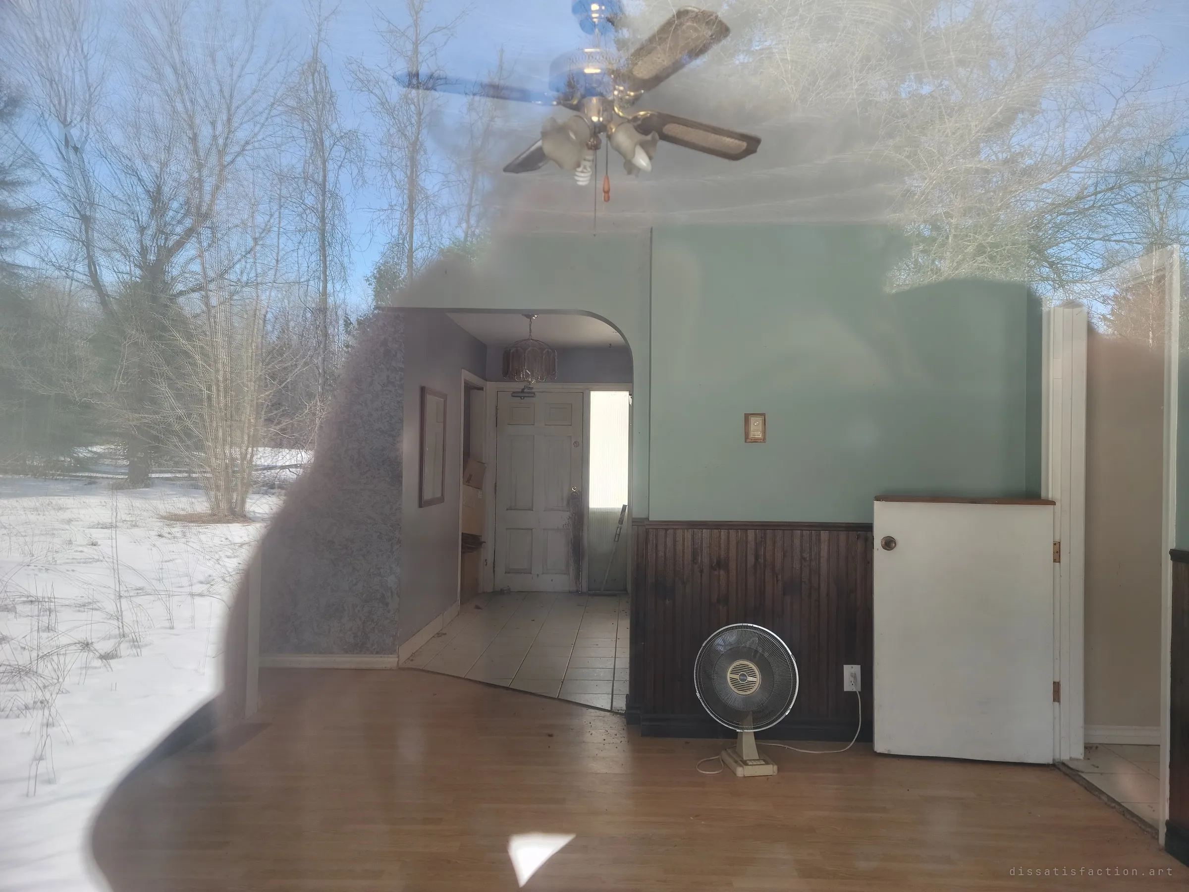 Looking through a smudged window into an empty mint-green room with a ceiling fan and a lone desk fan, the snowy woods reflected across the glass. The house stays sealed, seen only from outside.