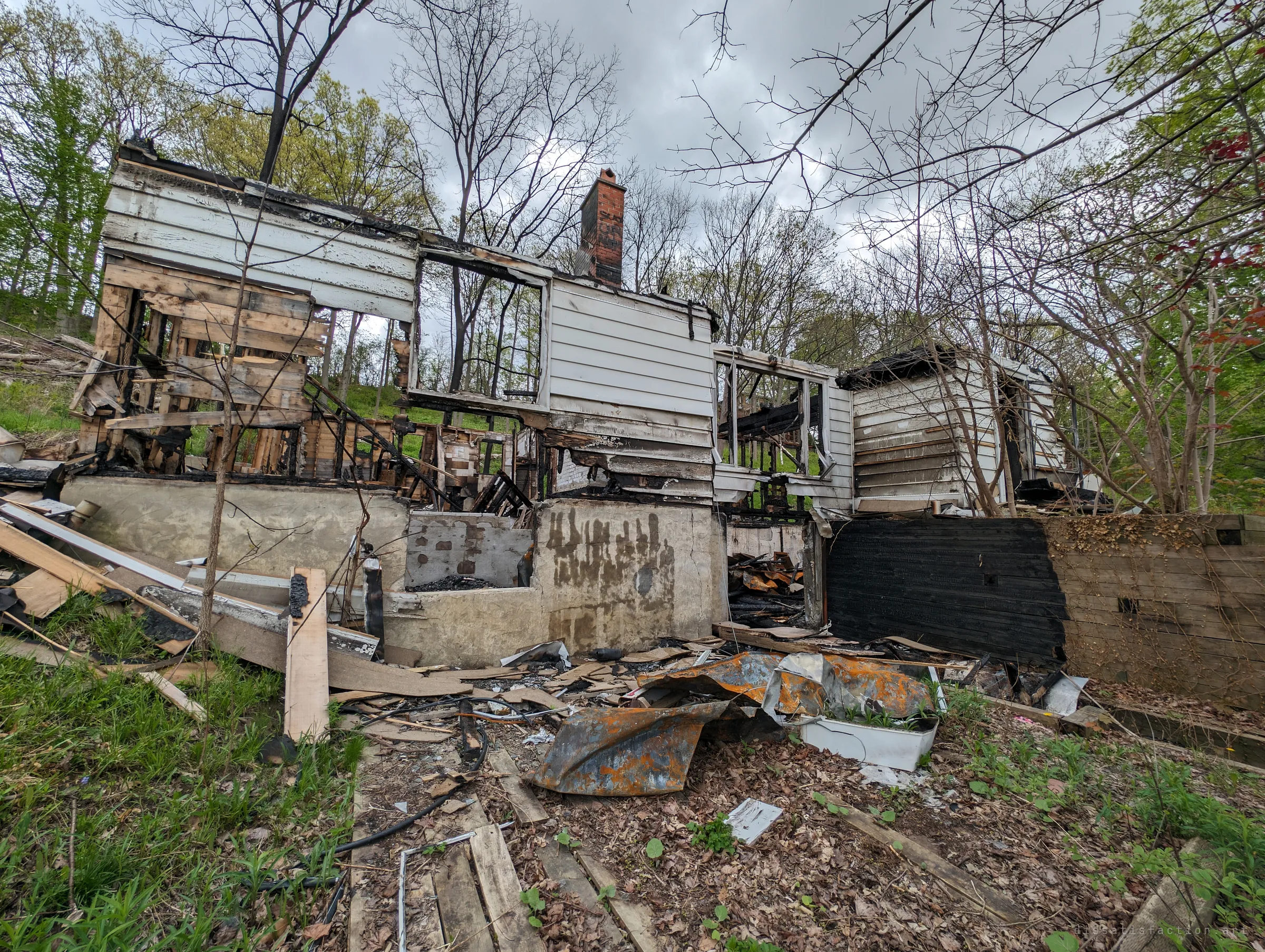 The white clapboard shell of a burned house on a wooded slope, its brick chimney still standing above a charred foundation and a yard strewn with scorched siding and rusted sheet metal. It sets the scene: the house is gutted but its outline still reads.