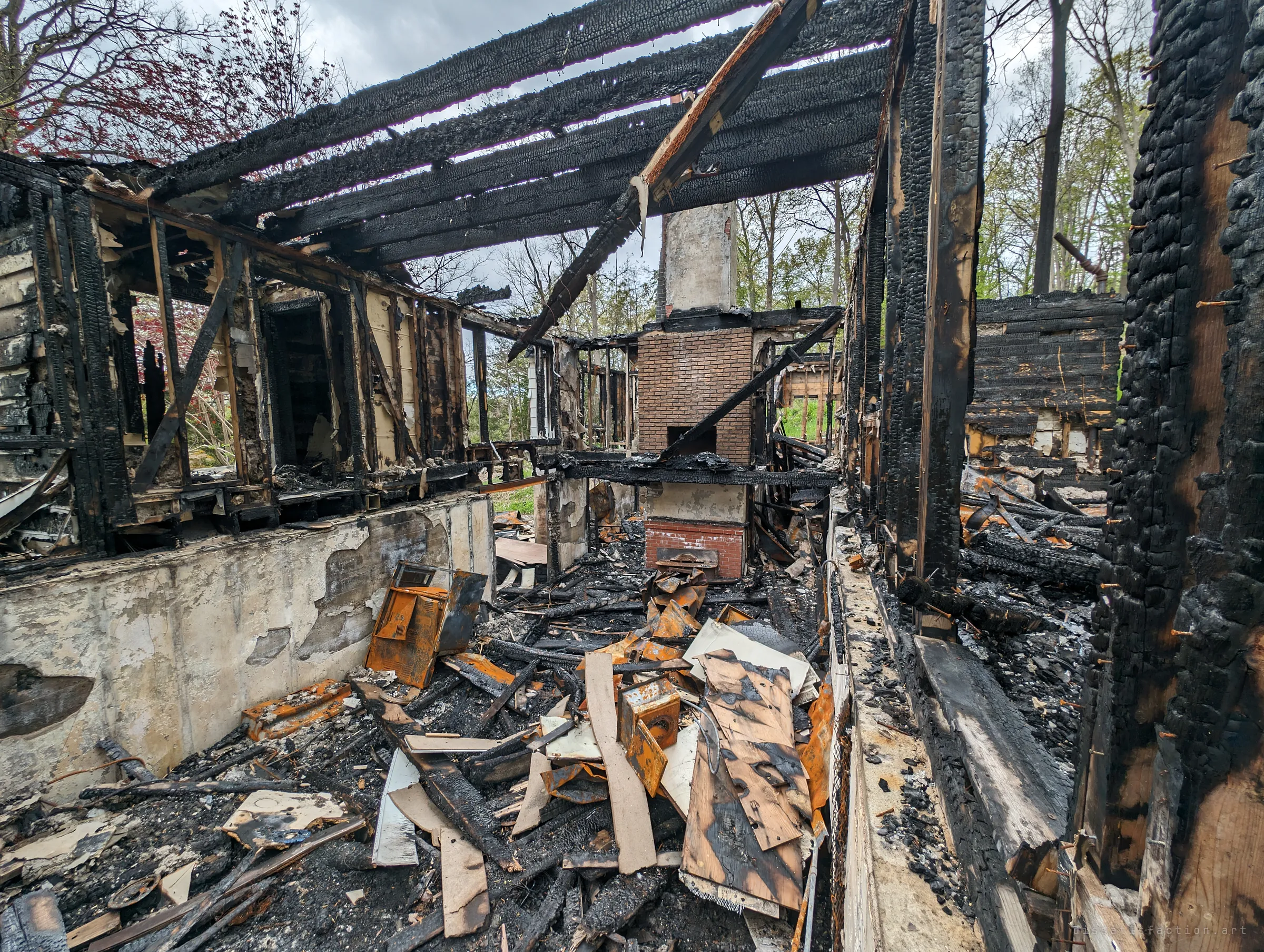 Inside the gutted house, blackened roof rafters cross open sky above a brick chimney and a floor buried in charred boards and heat-rusted appliance panels. The alligatored char on every beam shows how hot the fire burned.