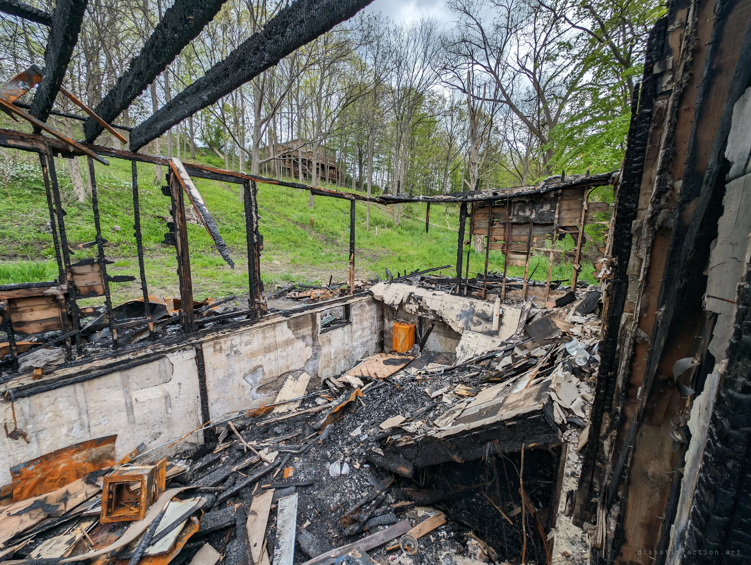 Looking out from the burned interior, charred wall studs frame a green hillside while the foundation below is filled with ash, collapsed flooring and a rusted metal cabinet. The empty window frames make the ruin feel like a skeleton.