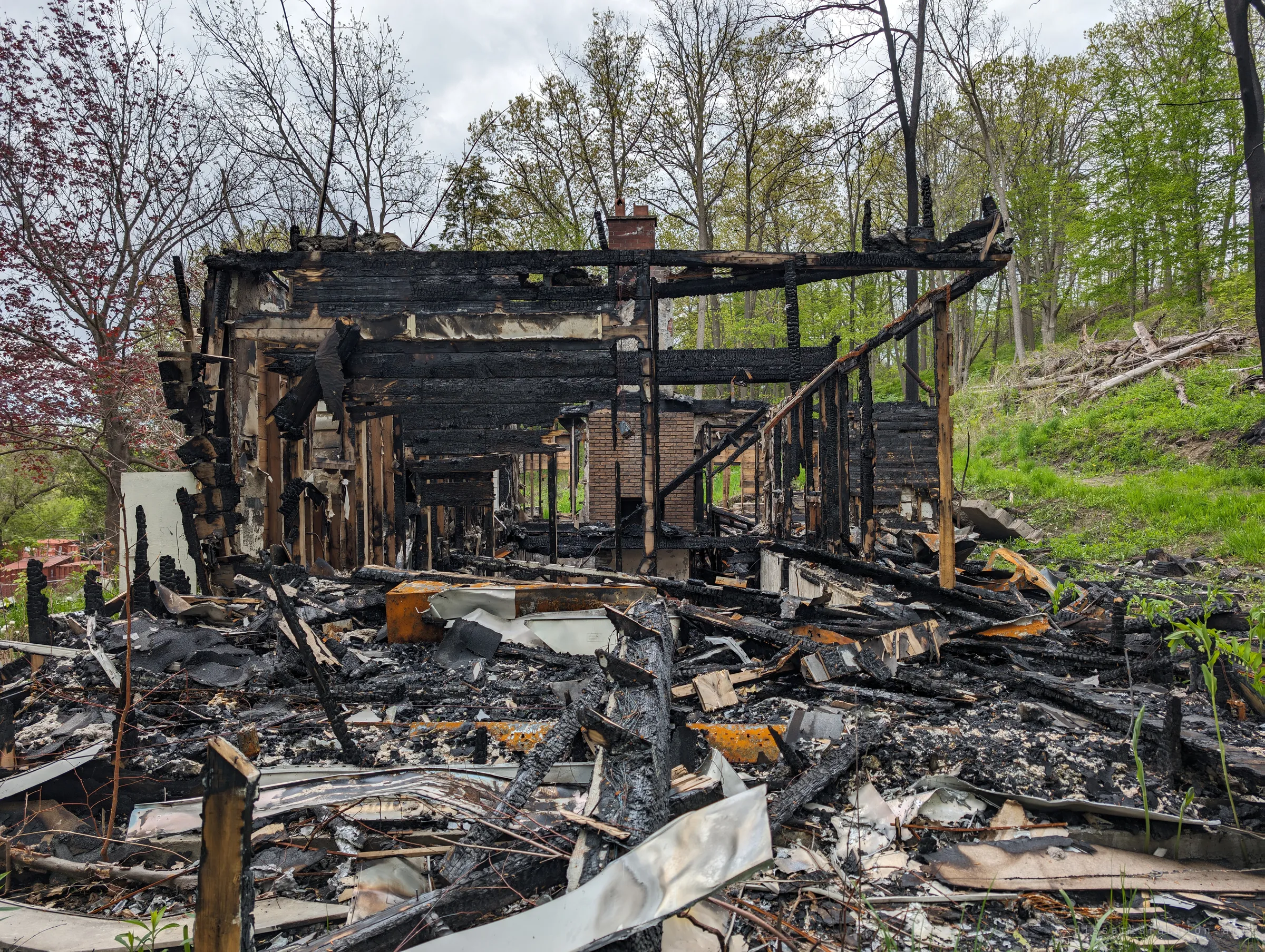 The collapsed side of the house seen from the rubble field, a skeleton of blackened framing with a brick chimney deep inside and a carpet of charred timber and twisted siding in front. It shows the full scale of what the fire left behind.
