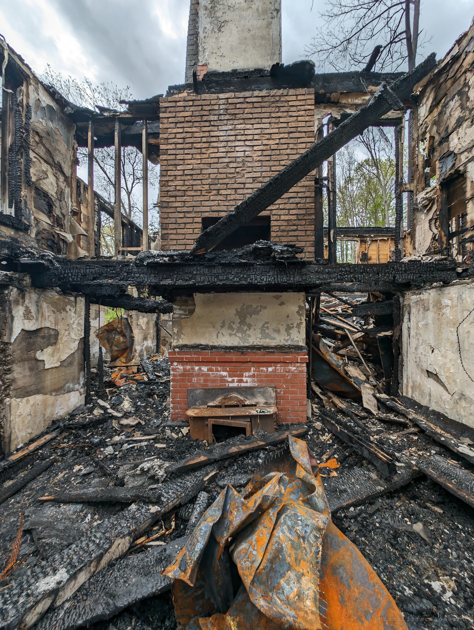 A two-storey brick chimney stands at the centre of the burned house, a fallen beam leaning across it and a small scorched bench by the hearth, with a crumpled rusted metal sheet in the foreground. The chimney is the one part of the house the fire could not take.