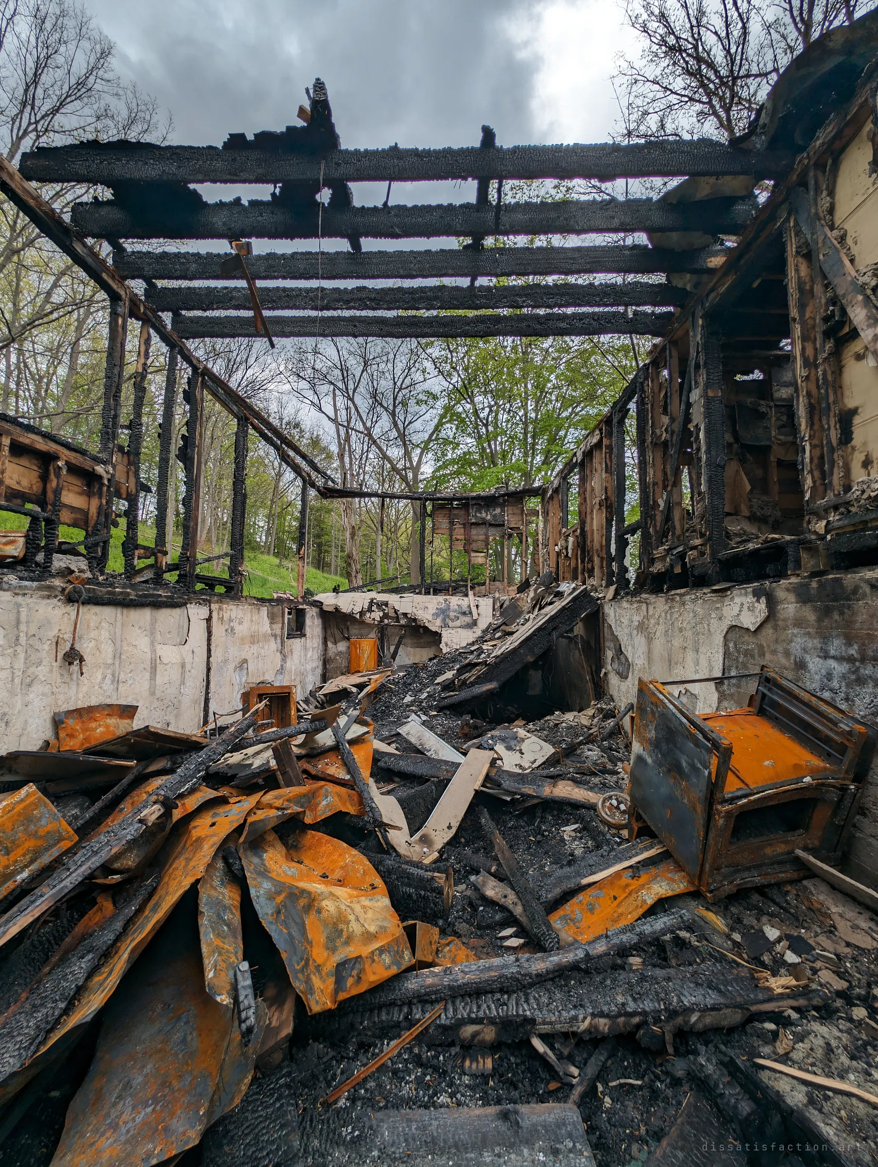 From the basement floor, roofless charred joists frame a stormy sky over walls of burned studs, while rusted appliances and warped metal sheets pile up in the foreground. The open roof turns the ruin into a frame for the trees beyond.