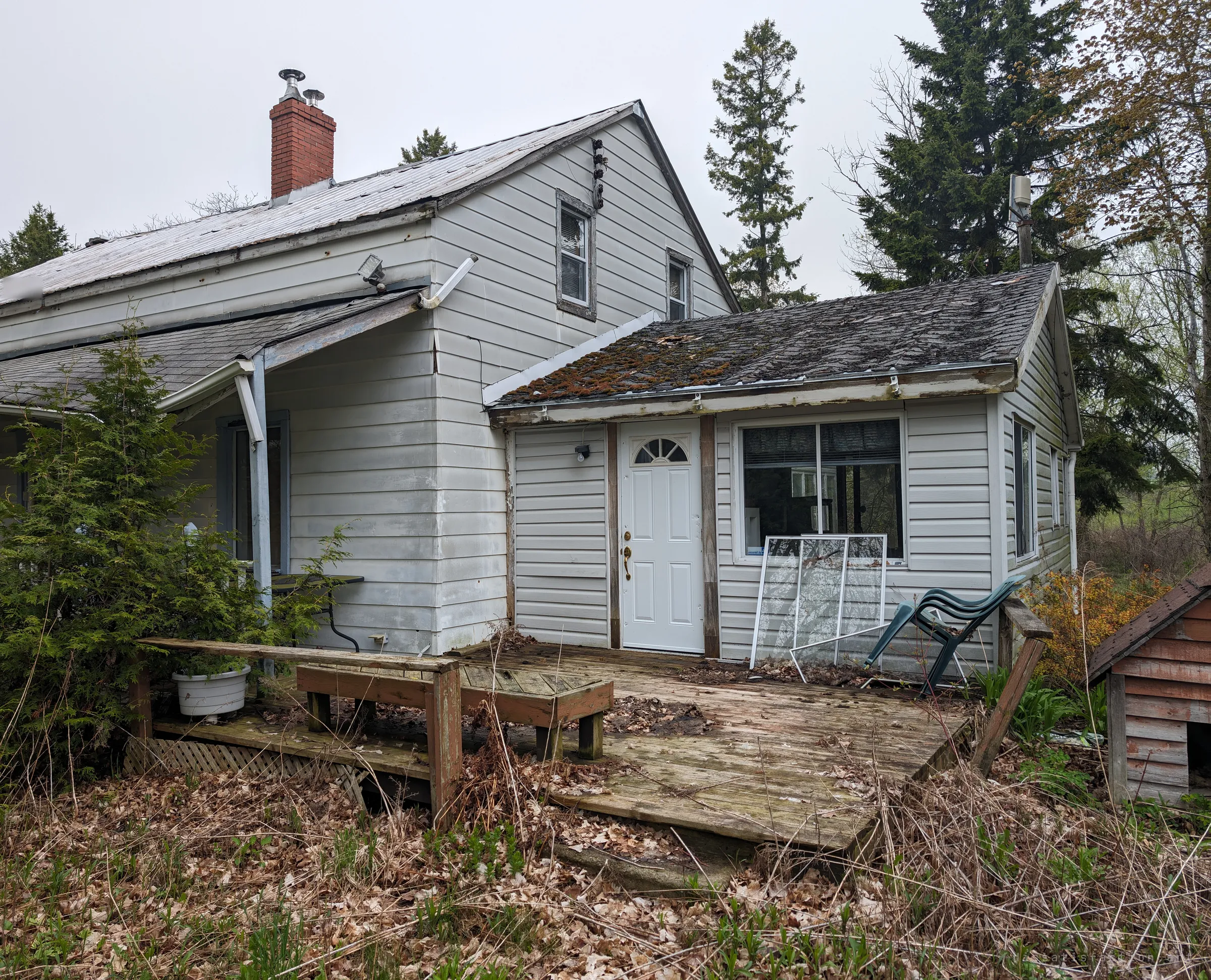 The side of the house with a moss-covered back addition, a rotting wooden deck and a toppled lawn chair. The rear shows how long the place has been left to weather.