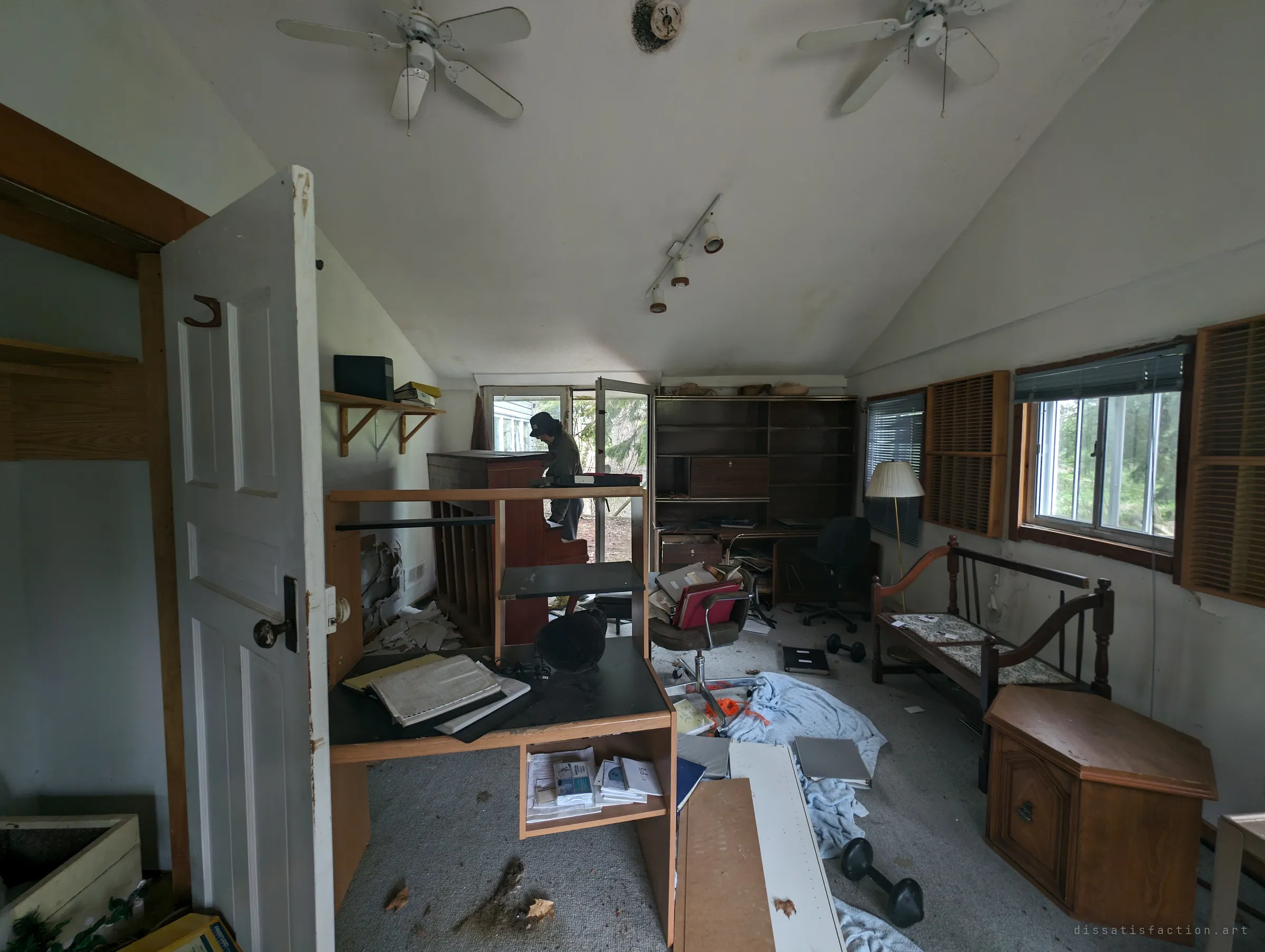 A vaulted study strewn with binders, papers and dumbbells, while a fellow explorer in a cap stands at an old upright piano by the open door. The room looks abandoned mid-task.