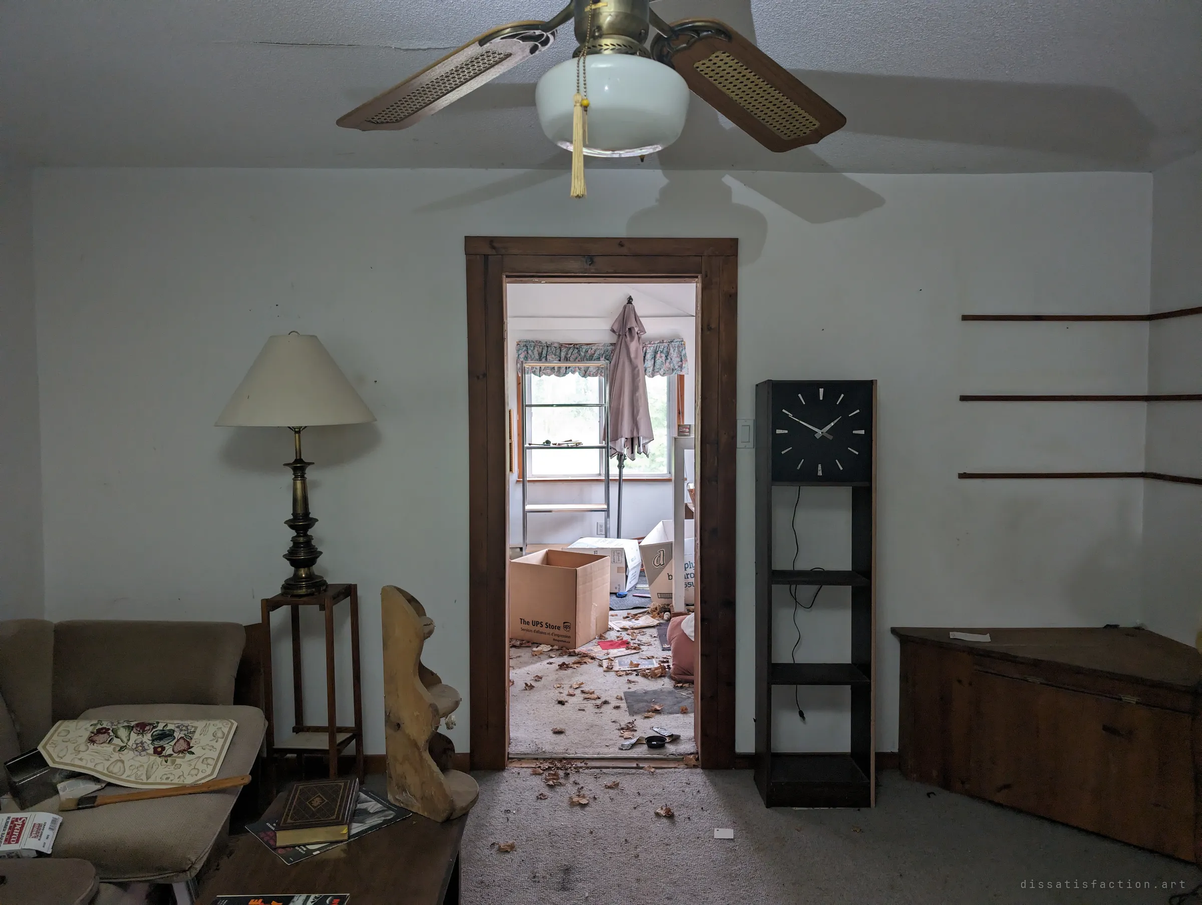 A living room with a brass lamp and a stopped wall clock, looking through a doorway into a bedroom littered with moving boxes and dead leaves. The house feels halfway packed up and then forgotten.