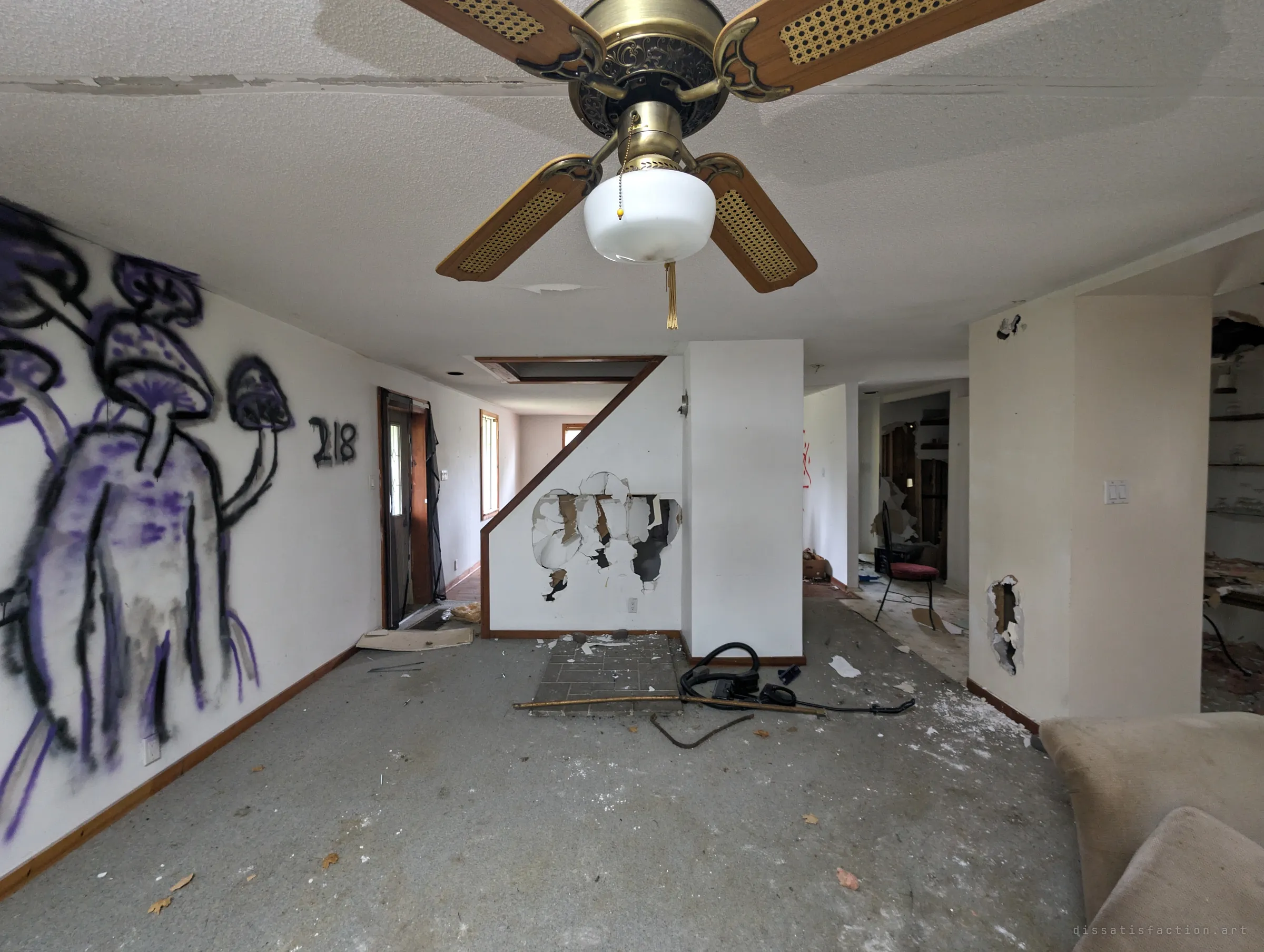 An open-plan living room under a cane-bladed ceiling fan, with purple mushroom graffiti across one wall and the drywall under the stairs smashed open. Vandals have clearly been through.