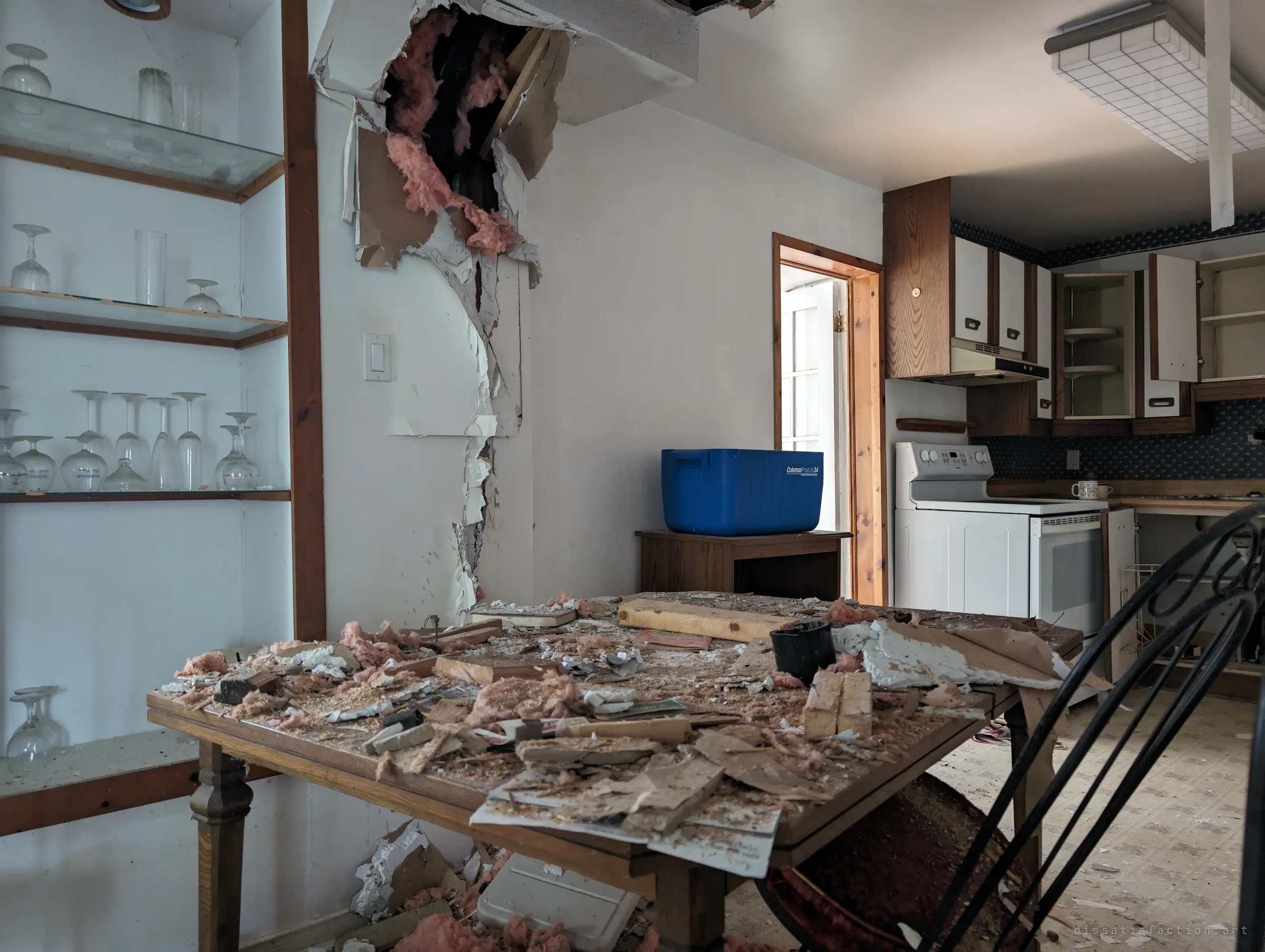A dining table buried in fallen insulation and drywall from a gaping hole in the ceiling, with a shelf of wine glasses still standing beside it and the kitchen beyond. The collapse sits right next to untouched glassware.